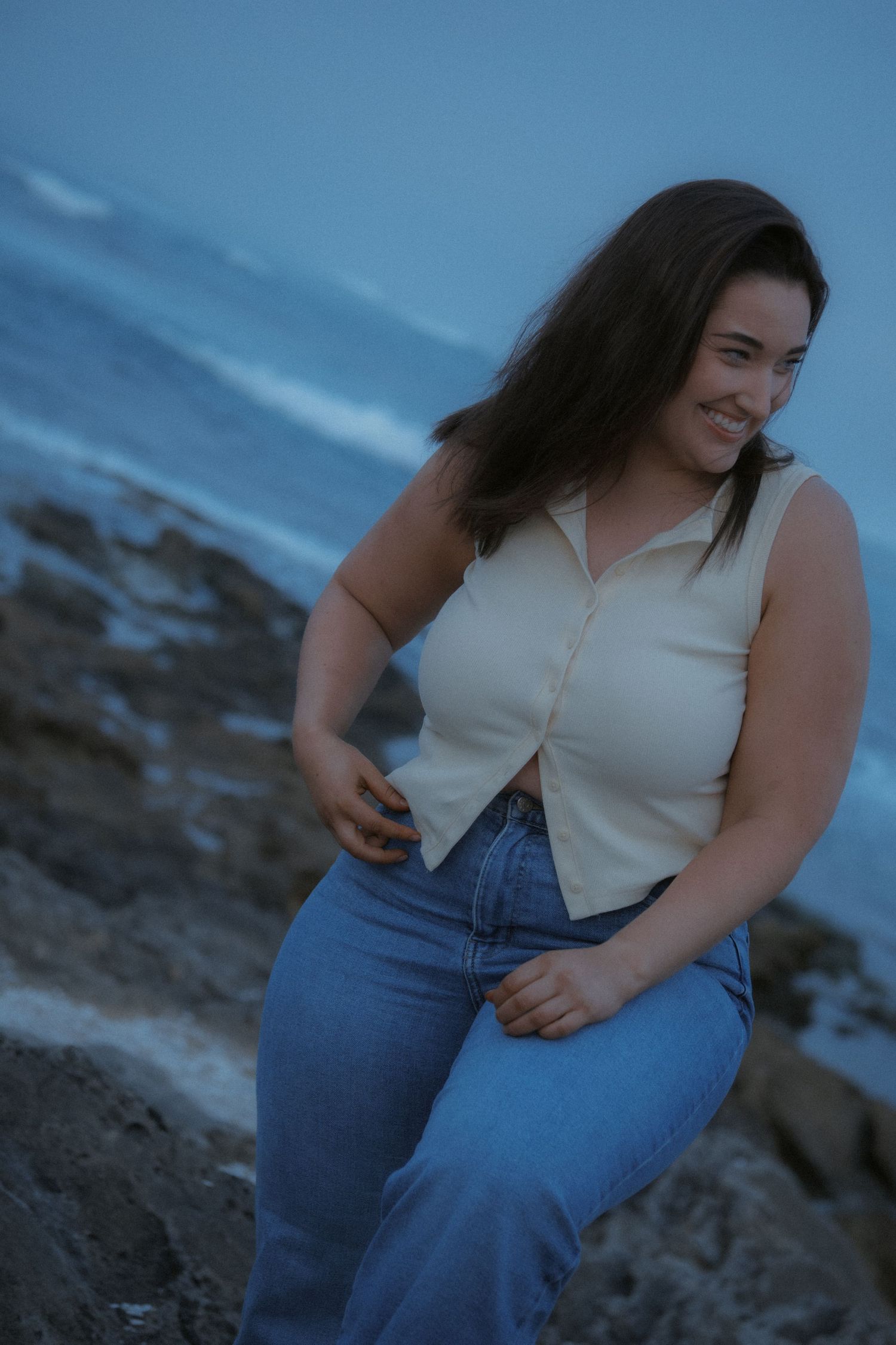 Young woman with long dark hair smiling on rocky coastal cliffs at dusk, wearing jeans and a cream top.