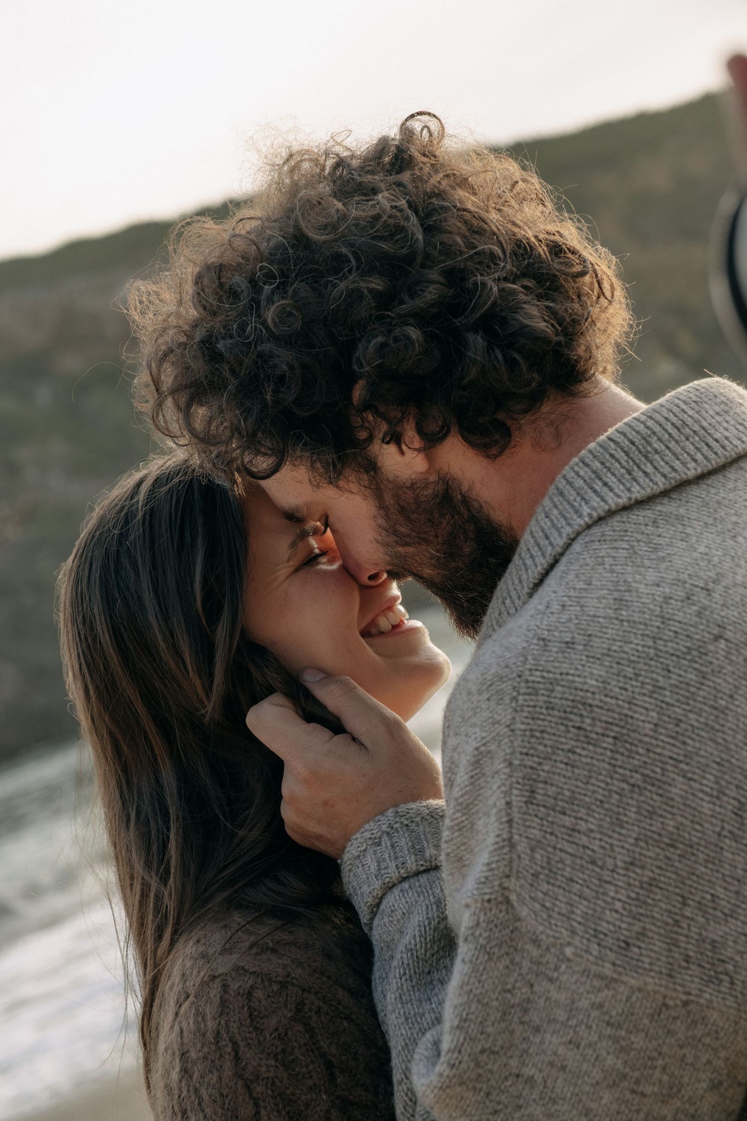 Bearded man with curly hair tenderly kissing a smiling woman on the forehead outdoors.