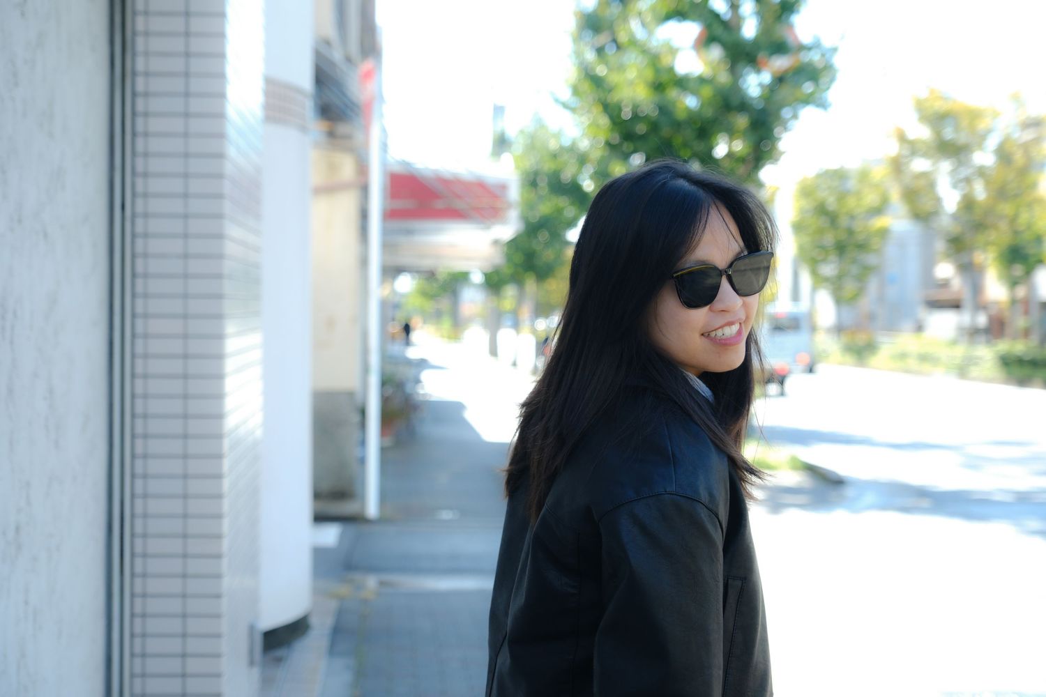 Stylish young woman with long black hair wearing sunglasses and a leather jacket, smiling on a sunny street.