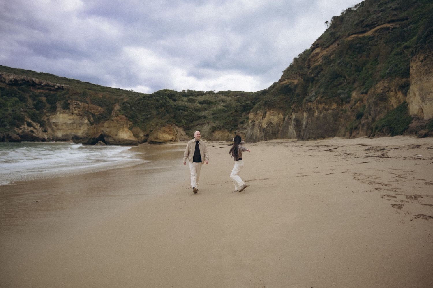 Two people walking hand in hand on a sandy beach with dramatic cliffs and cloudy skies in the background.