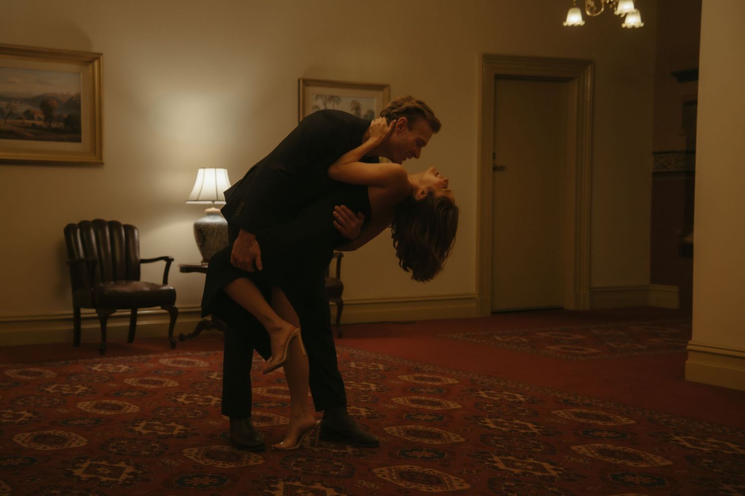 A couple shares a romantic dip and kiss while dancing in a dimly lit room with a red patterned carpet.