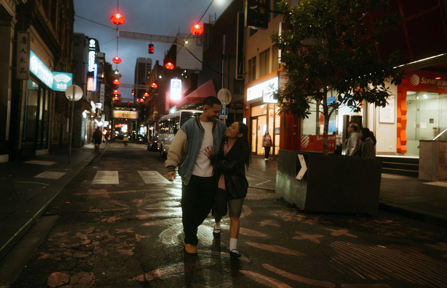 Couple walking hand in hand on a wet Chinatown street at night, illuminated by red lanterns and shop lights.