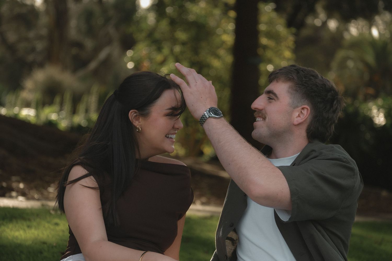 A smiling couple shares a playful moment outdoors in a lush green park, with the man gently touching the woman's hair.
