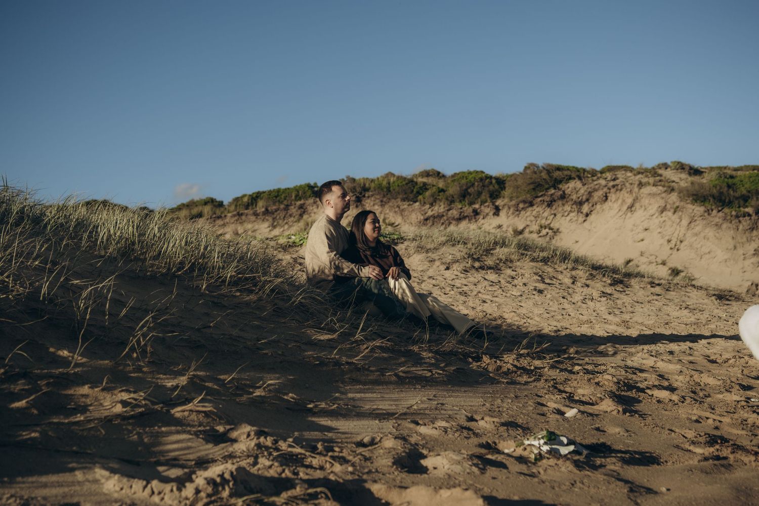 Couple sitting together on sandy beach dunes with green grass and clear blue sky in background.