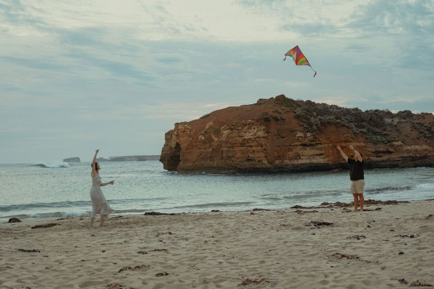 Couple flying a colorful kite on a scenic beach with rocky coastal cliffs nearby.