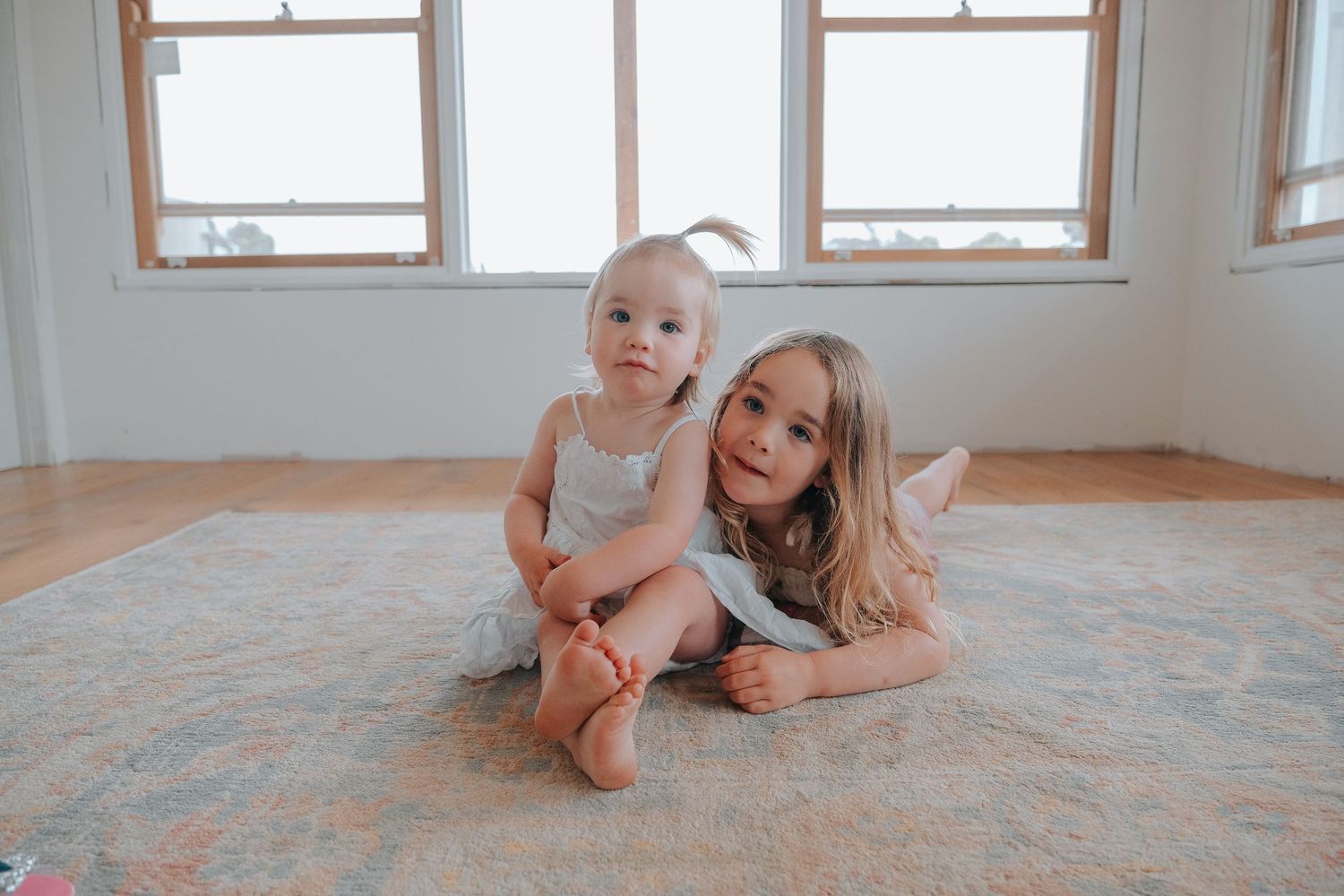Two young sisters sit together on a rug in a bright room with large windows, smiling at the camera.