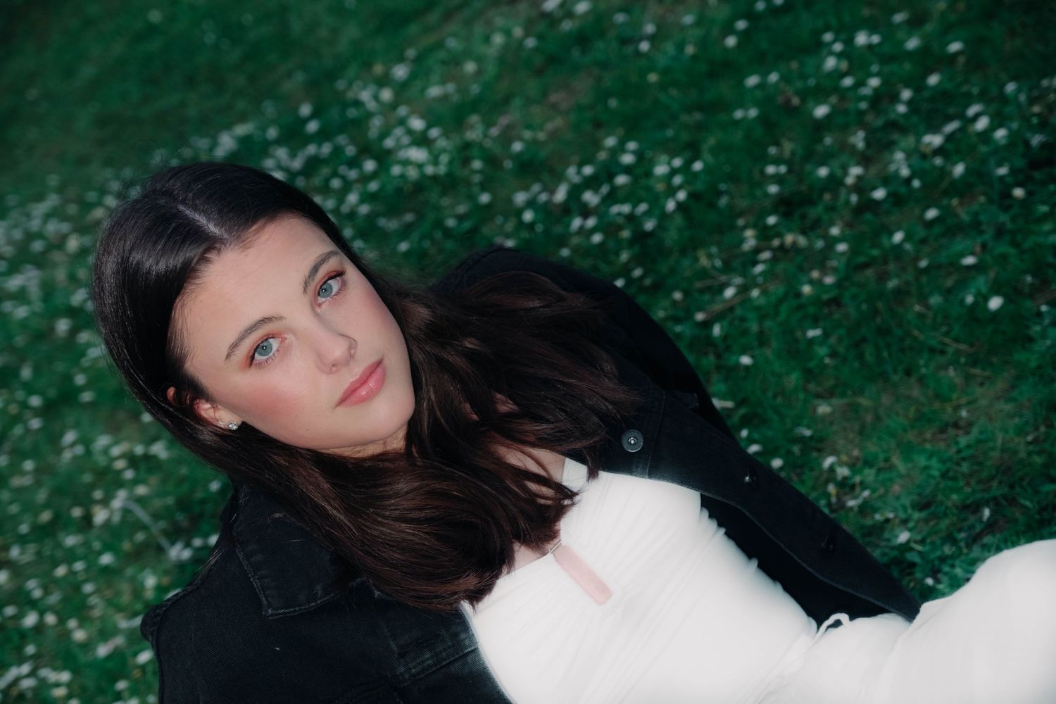 Beautiful young brunette woman lying on a flower-dotted green lawn wearing a black jacket.