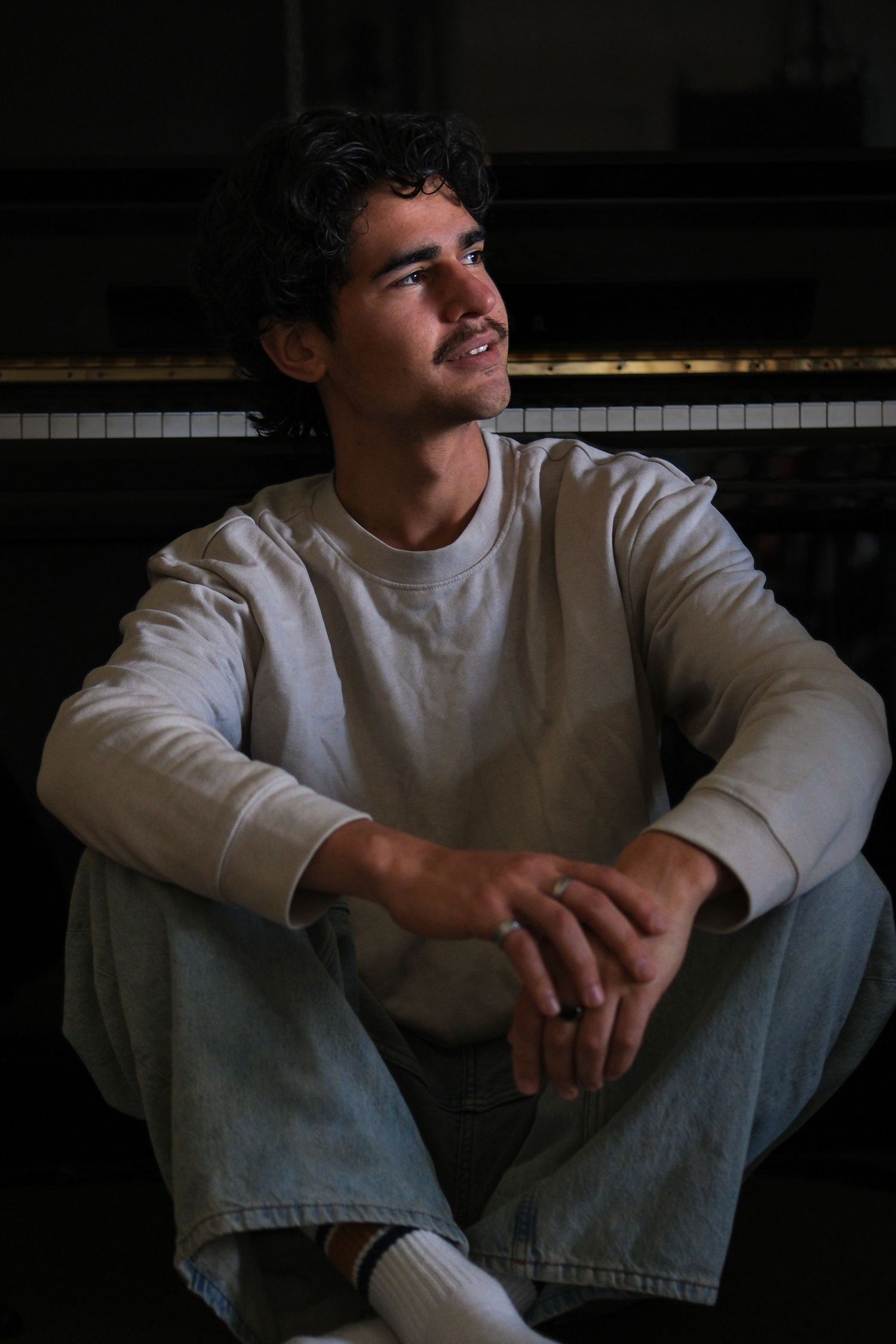 Young man with curly hair sitting on floor leaning against piano in dim light, documentary style photography Visuals by Dani.