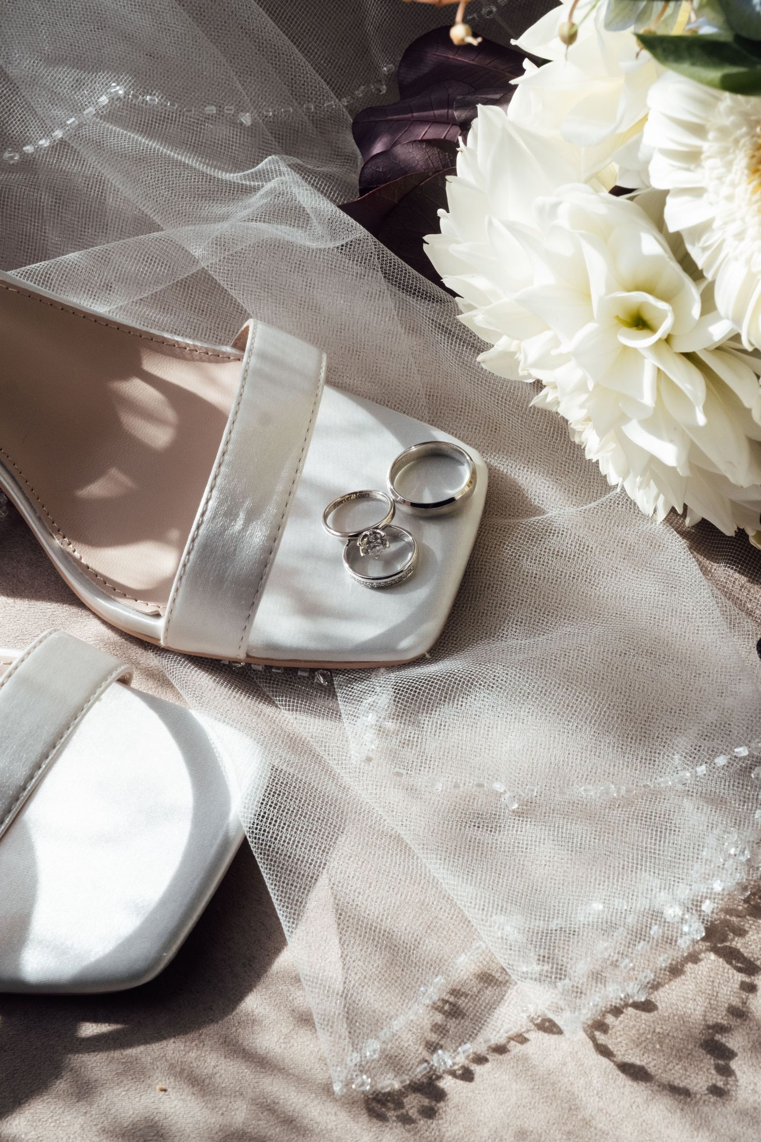Elegant wedding flat lay with white heels, silver rings, bridal veil, clutch, and white floral bouquet on neutral background.