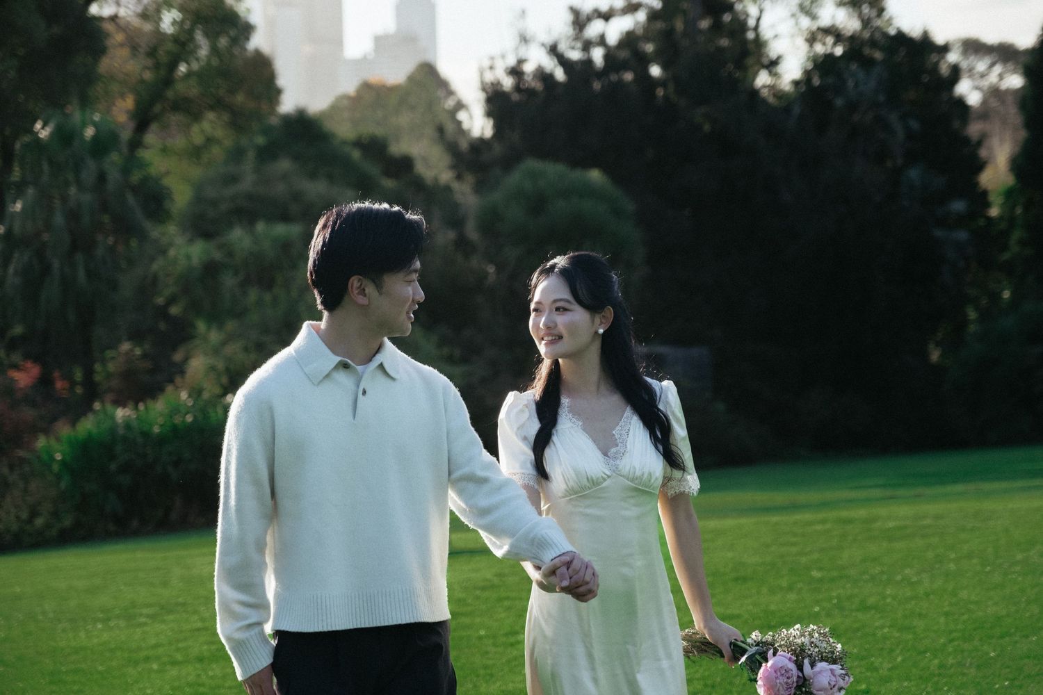 A young couple walks hand-in-hand on a green lawn, the woman in a white dress holding flowers, the man in a cream sweater.