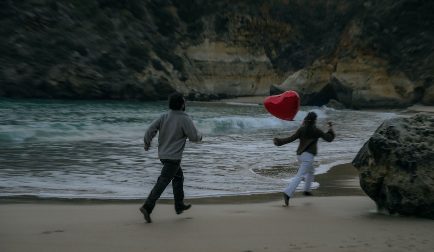 Couple running on a beach, one holding a red heart-shaped balloon near rocky cliffs.