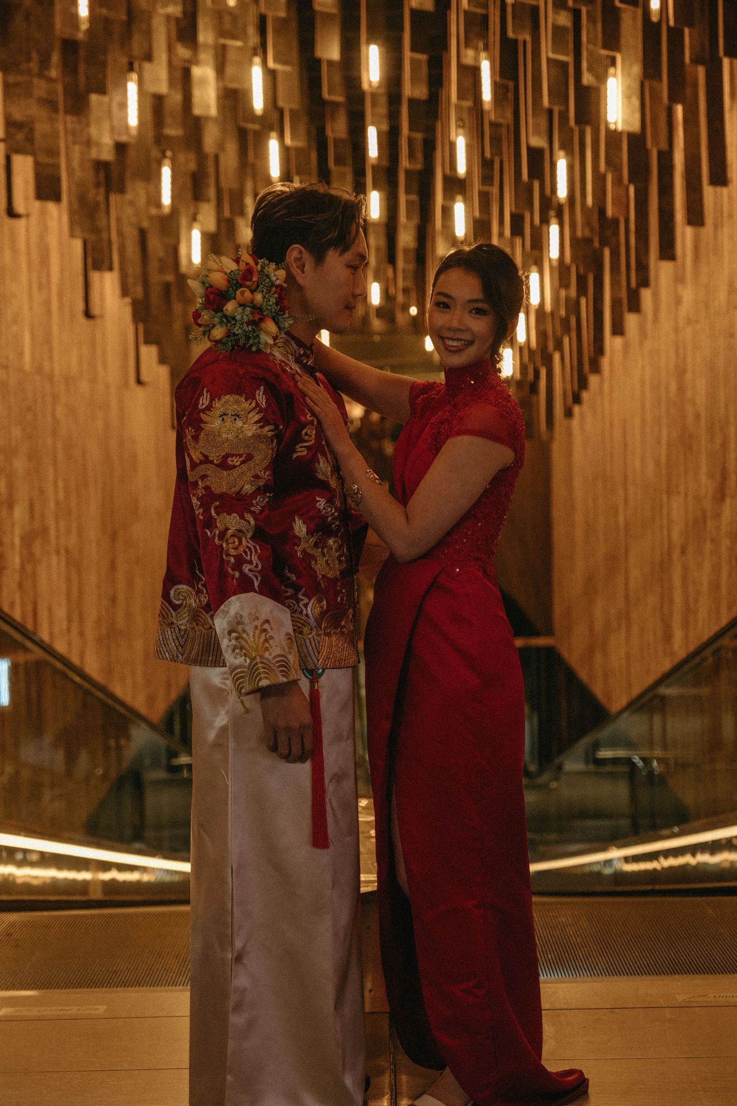 Couple in traditional Chinese wedding attire, groom in red dragon robe, bride in red qipao, posing in elegant lit venue.