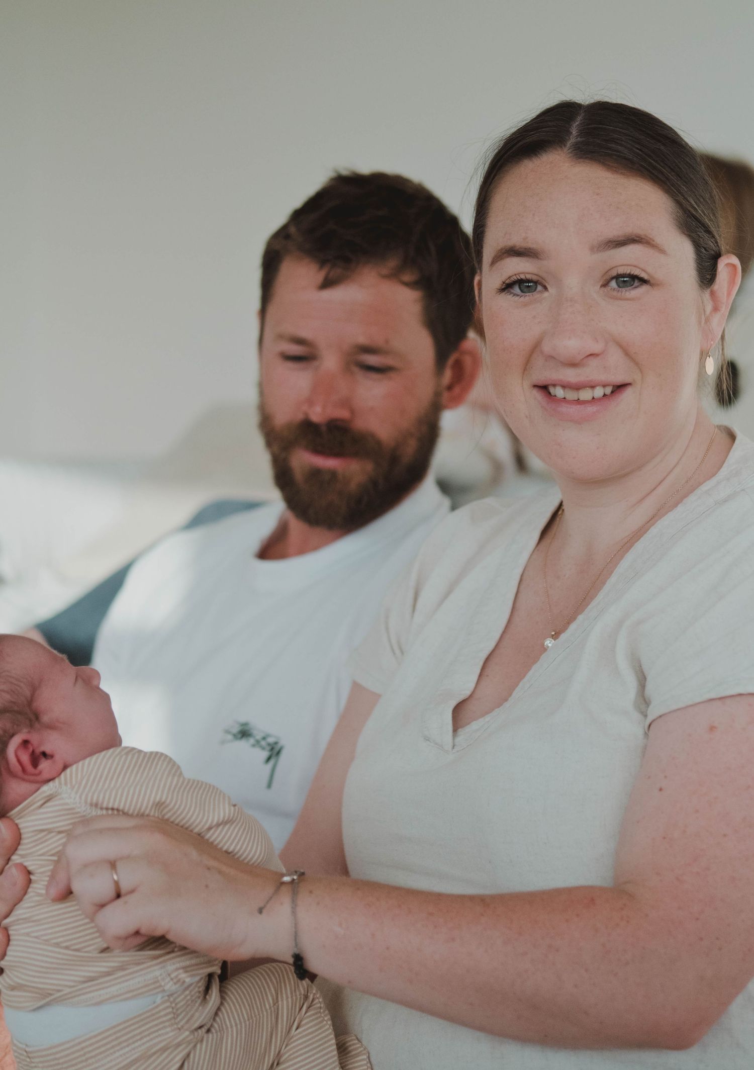 Smiling mother holds newborn baby while father looks on lovingly in a bright, cozy home setting.