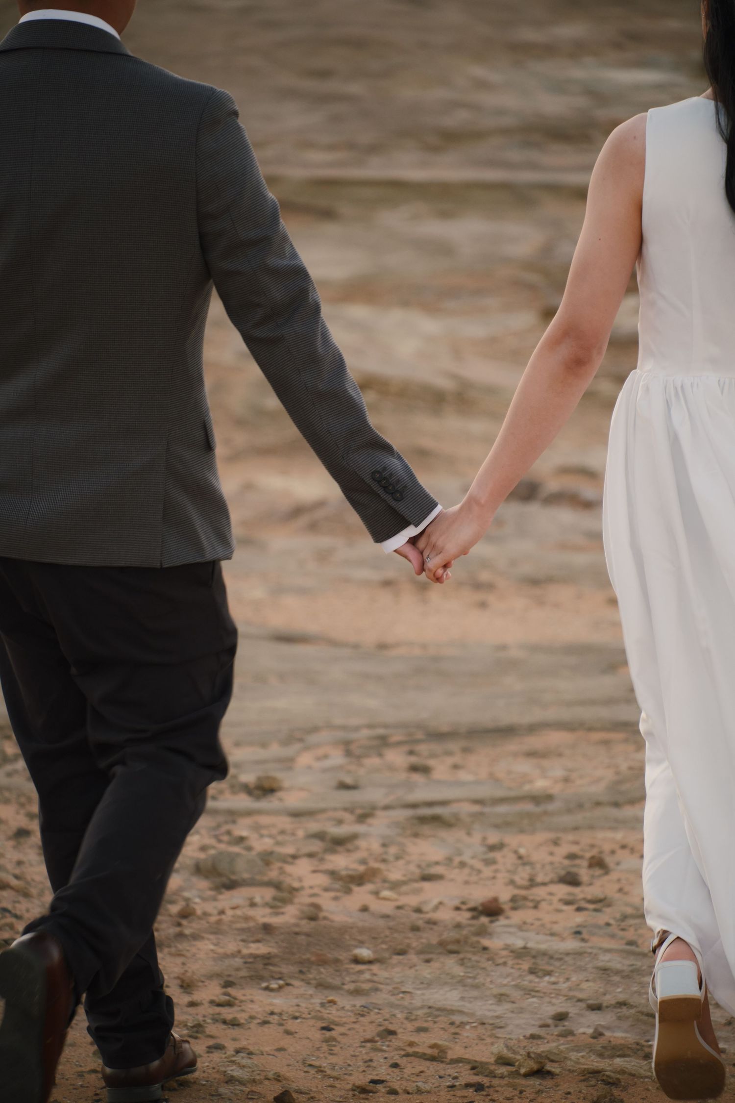 Bride and groom holding hands walking on sandy terrain, rear view of wedding couple in white dress and gray suit.