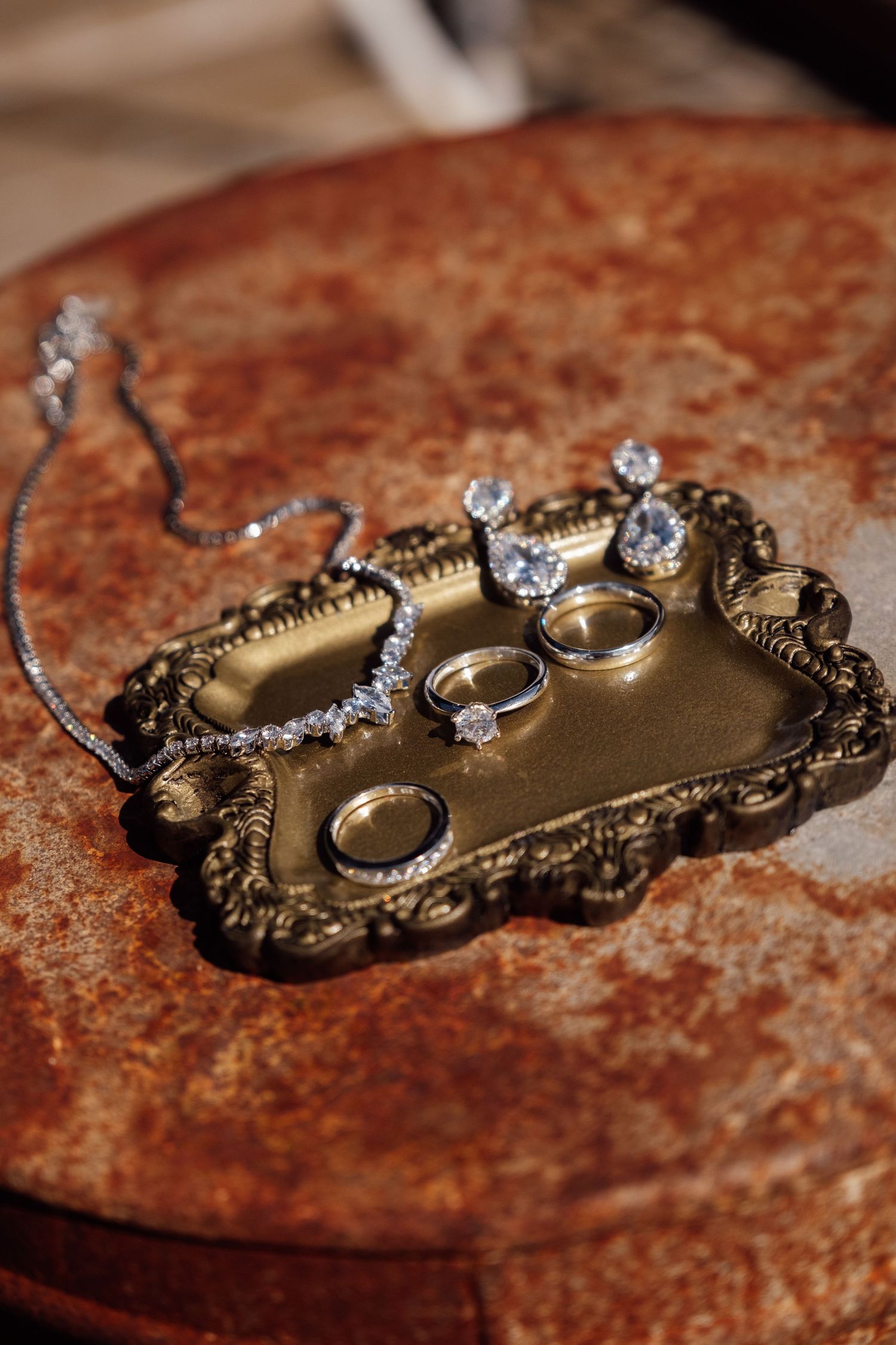 Elegant wedding rings and diamond jewelry displayed on ornate gold tray with necklace on rustic surface.