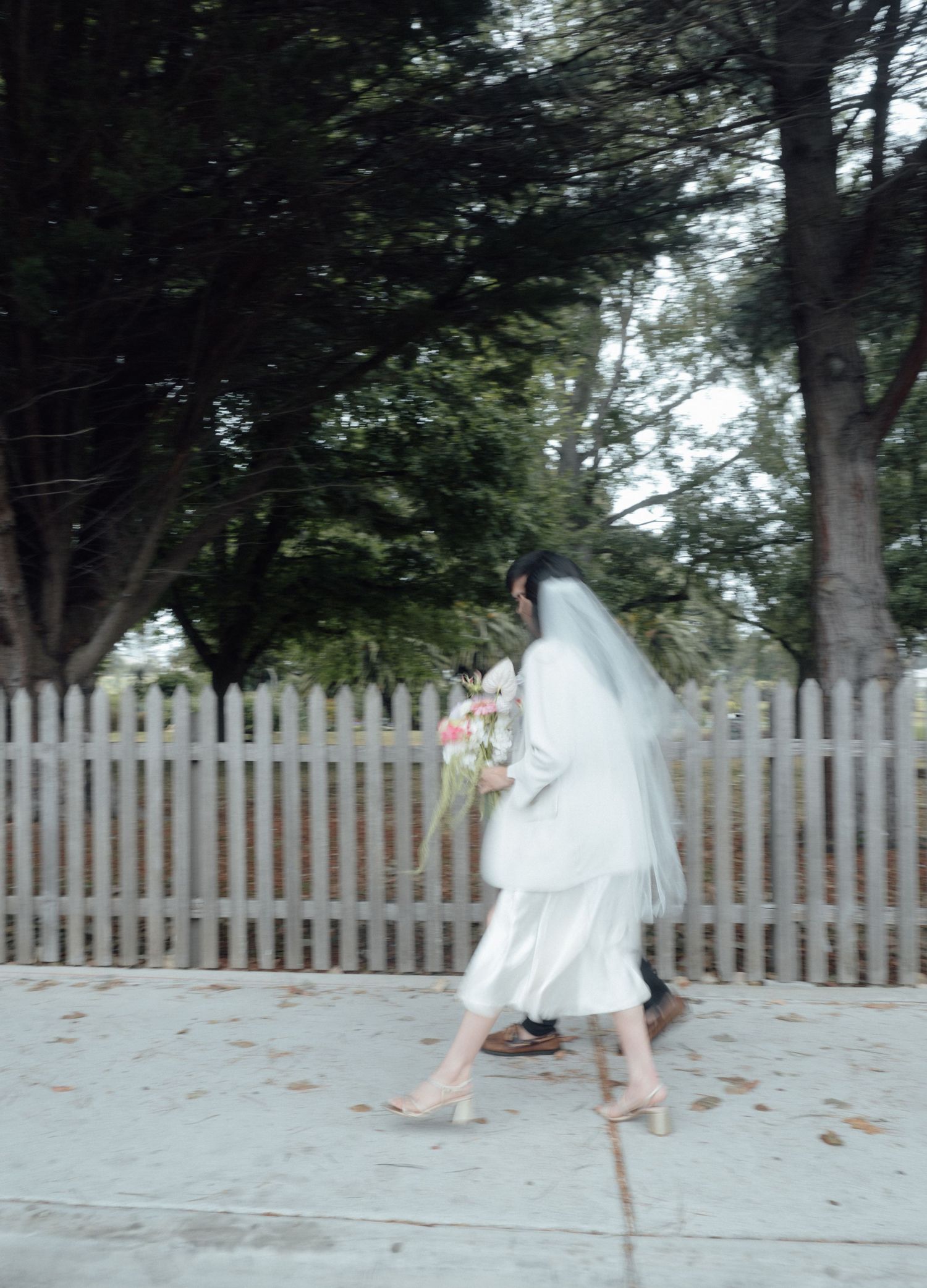 A bride in a white wedding dress and veil walks along a sidewalk holding a floral bouquet near a white picket fence.