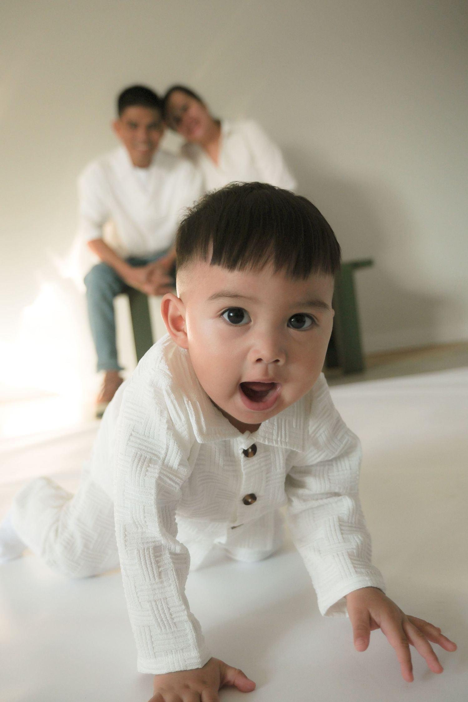 Curious baby in white outfit crawling toward camera with parents sitting behind.