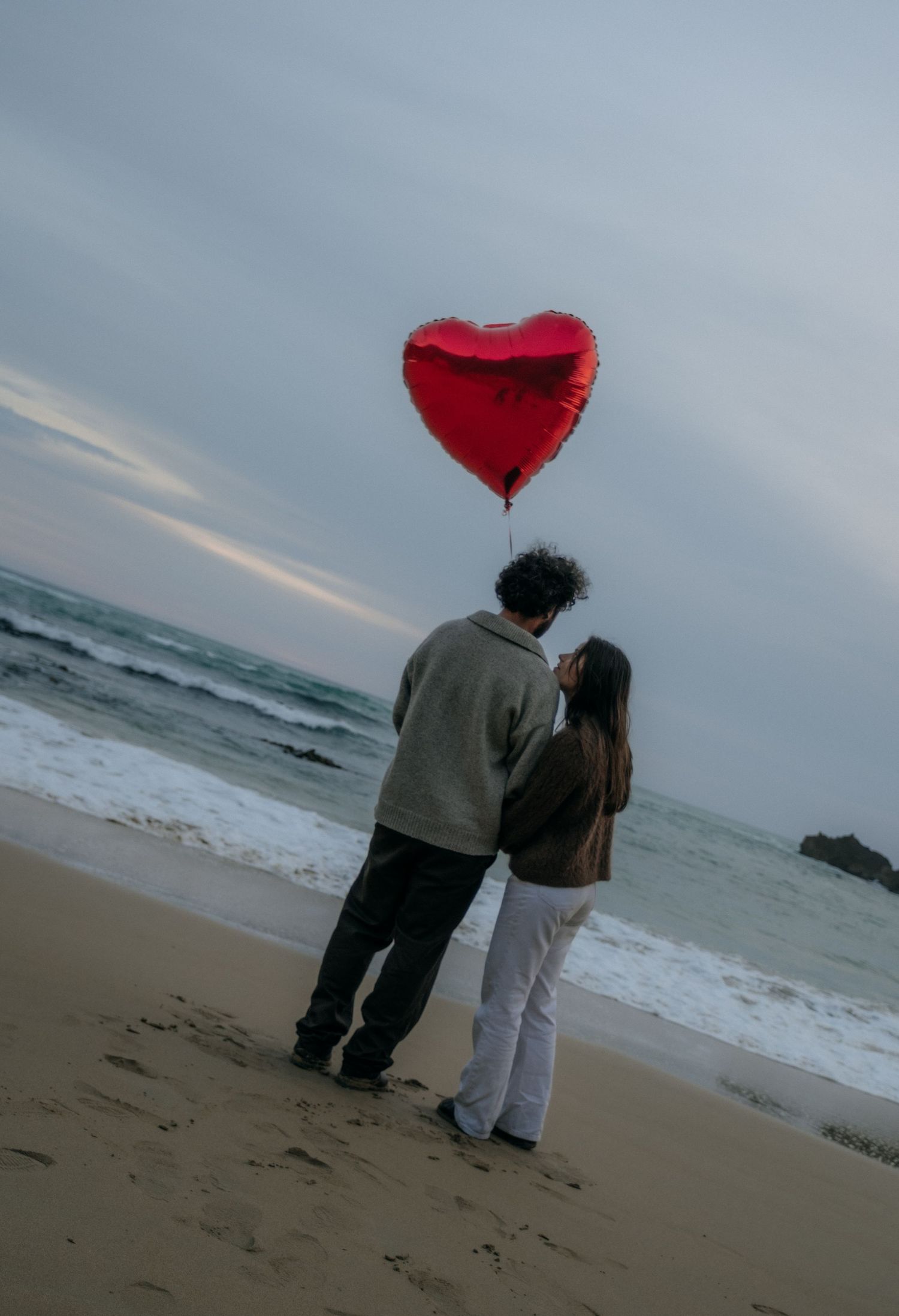 Romantic couple standing on a beach holding a red heart balloon at dusk by the ocean. Great Ocean Road photography.