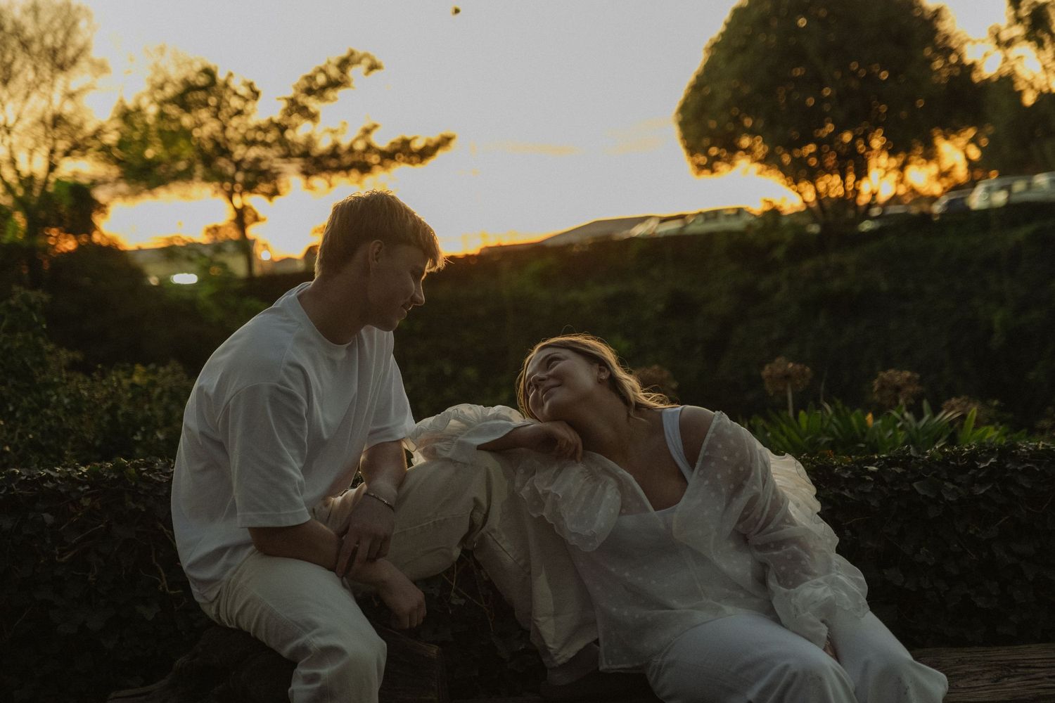 Couple in white outfits laughing together at sunset in a lush garden setting.
