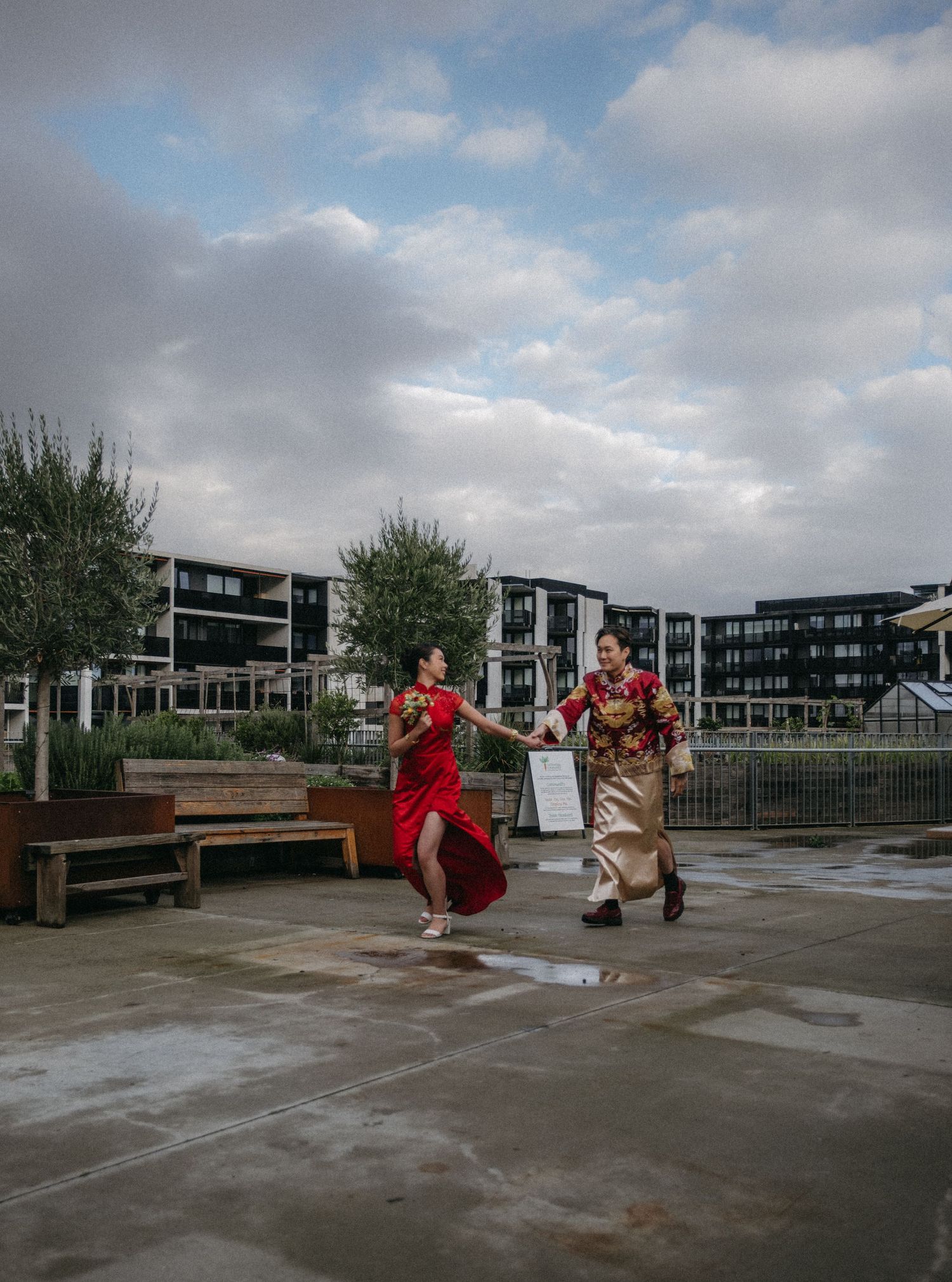Couple in traditional Chinese attire holding hands and running outdoors near modern buildings under a cloudy sky.
