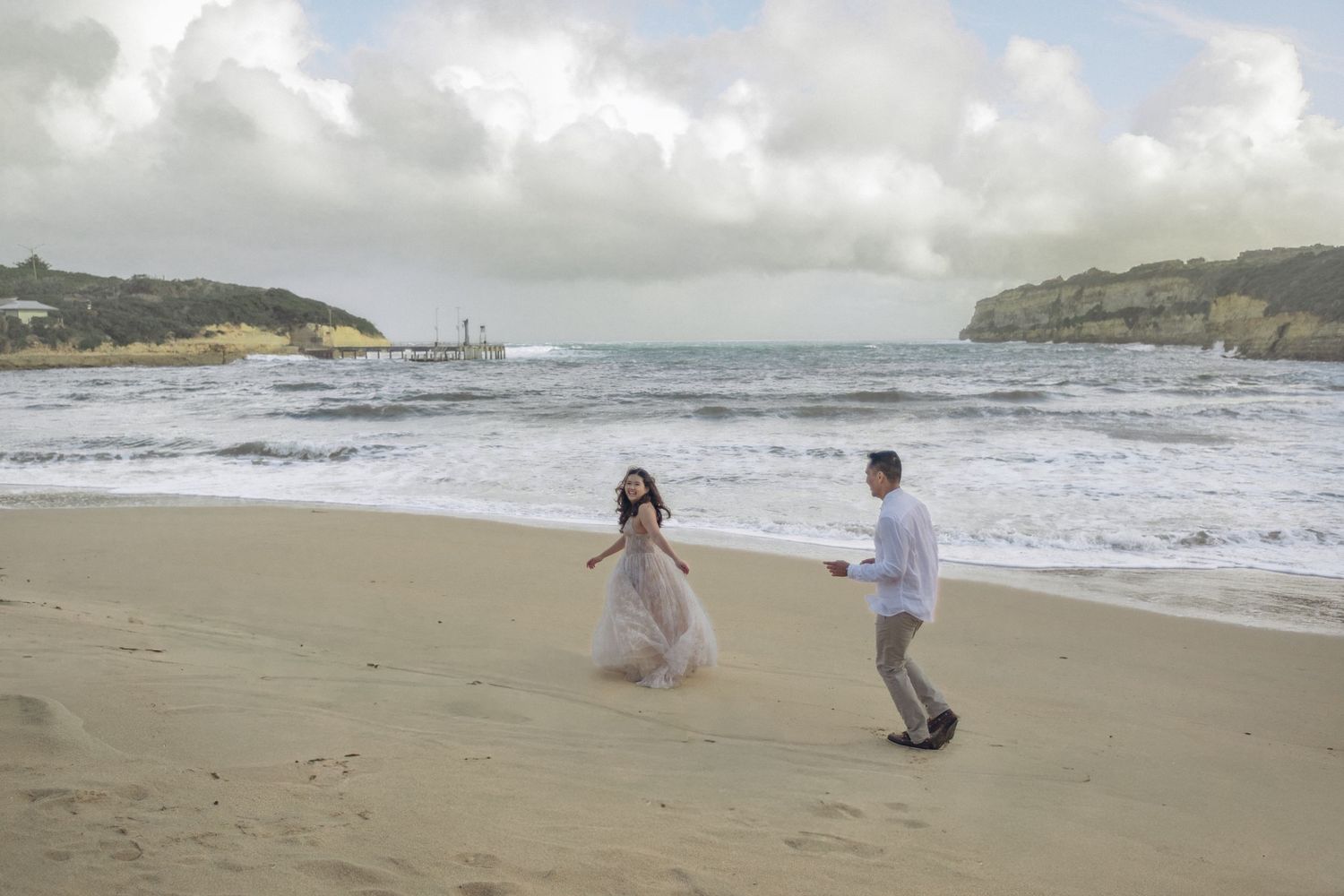 Couple in wedding attire walking on a sandy beach with ocean waves and cloudy skies in the background.