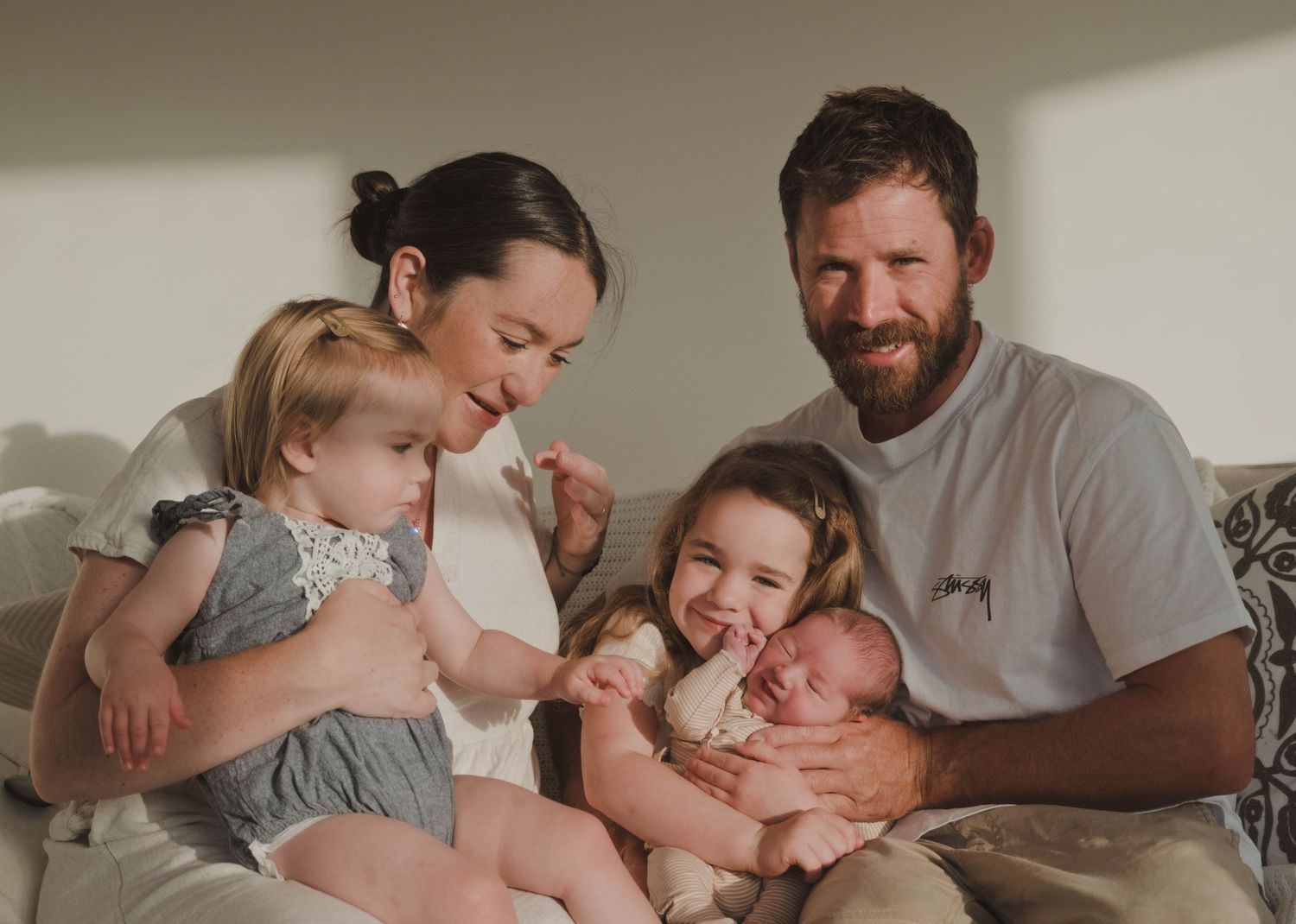 Happy family of five with two young daughters and a newborn baby sitting together on a couch.