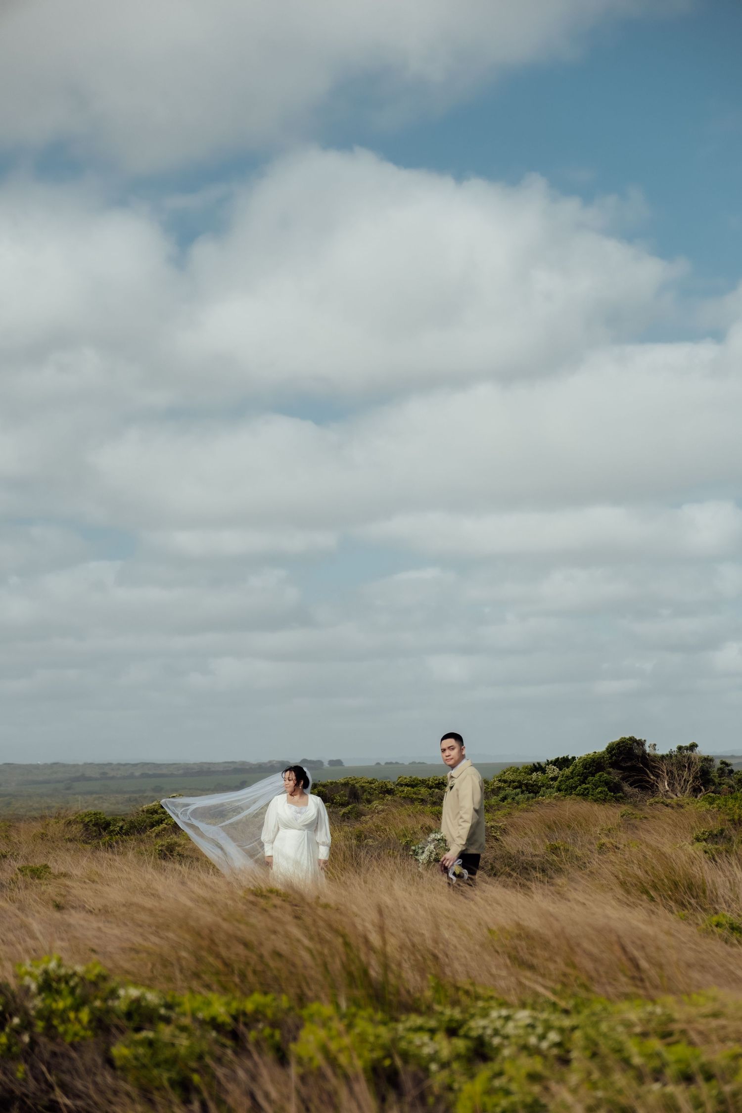 Bride and groom stand in windswept coastal grasslands, her veil flowing in the breeze under a dramatic cloudy sky.