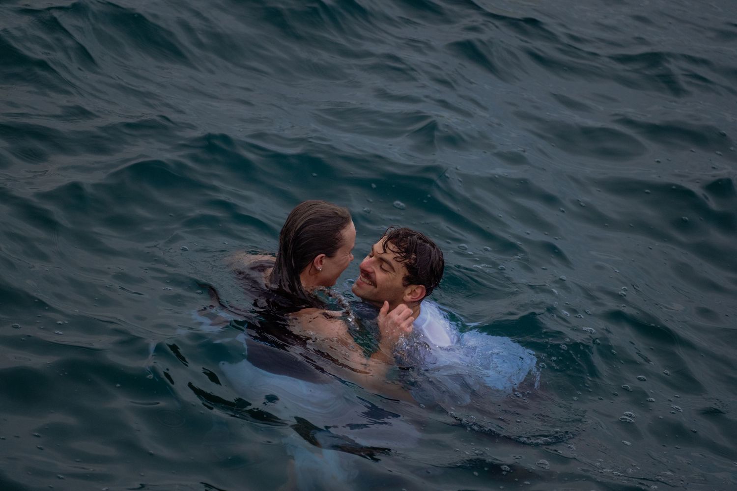 Two people embracing in open ocean water, staying afloat together in calm blue-gray sea waves.