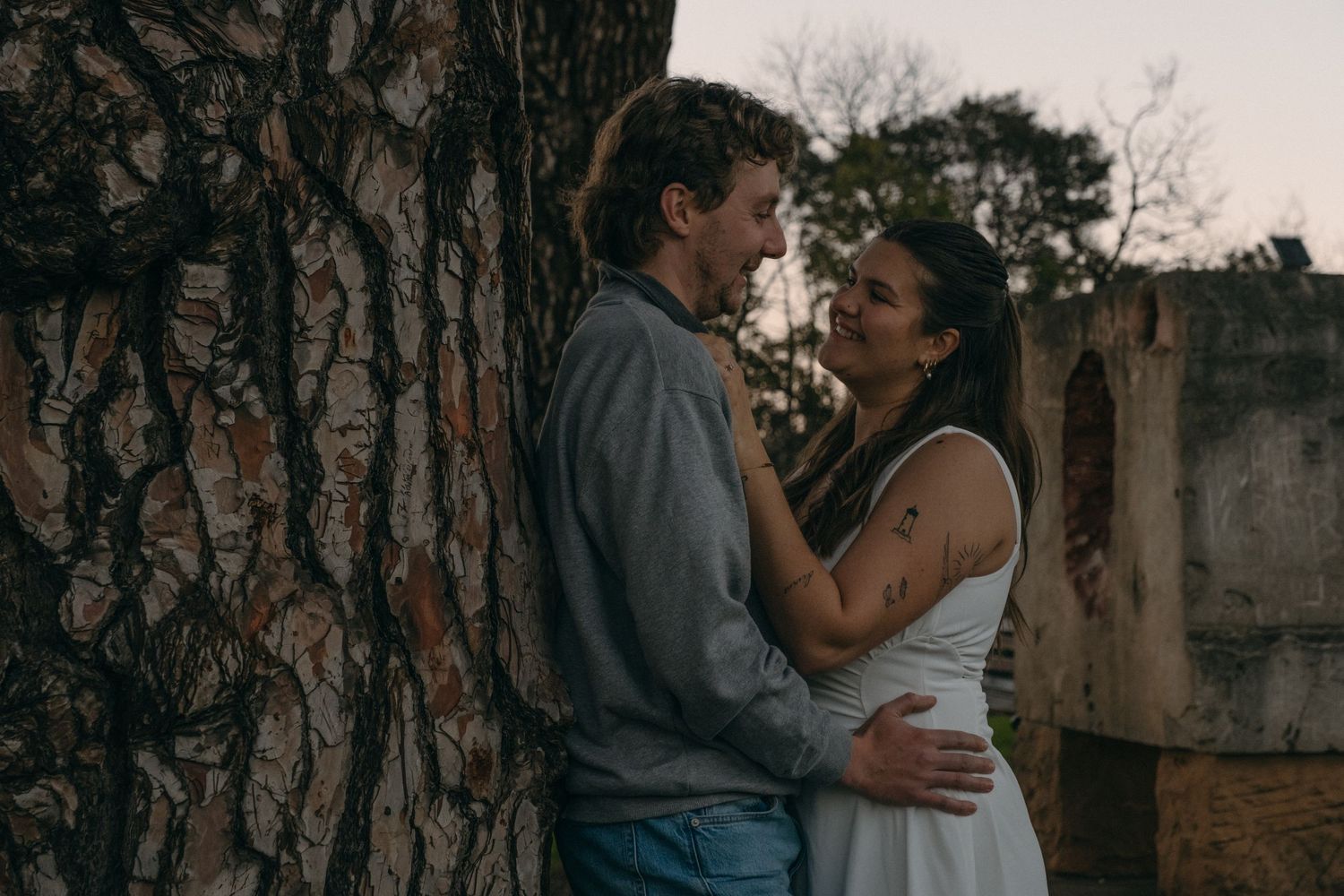 Couple embracing romantically beside a large tree trunk outdoors at dusk, woman in white dress with floral tattoo.