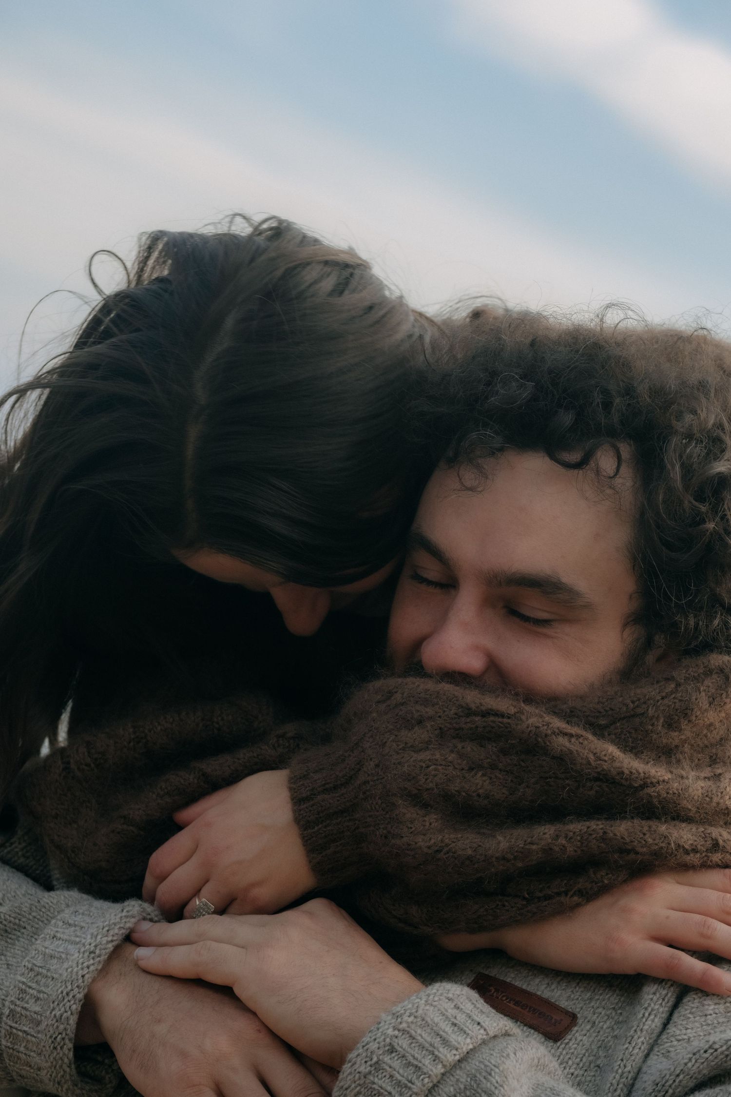 Couple embracing tenderly outdoors, woman with dark hair hugging man in brown sweater against cloudy sky.