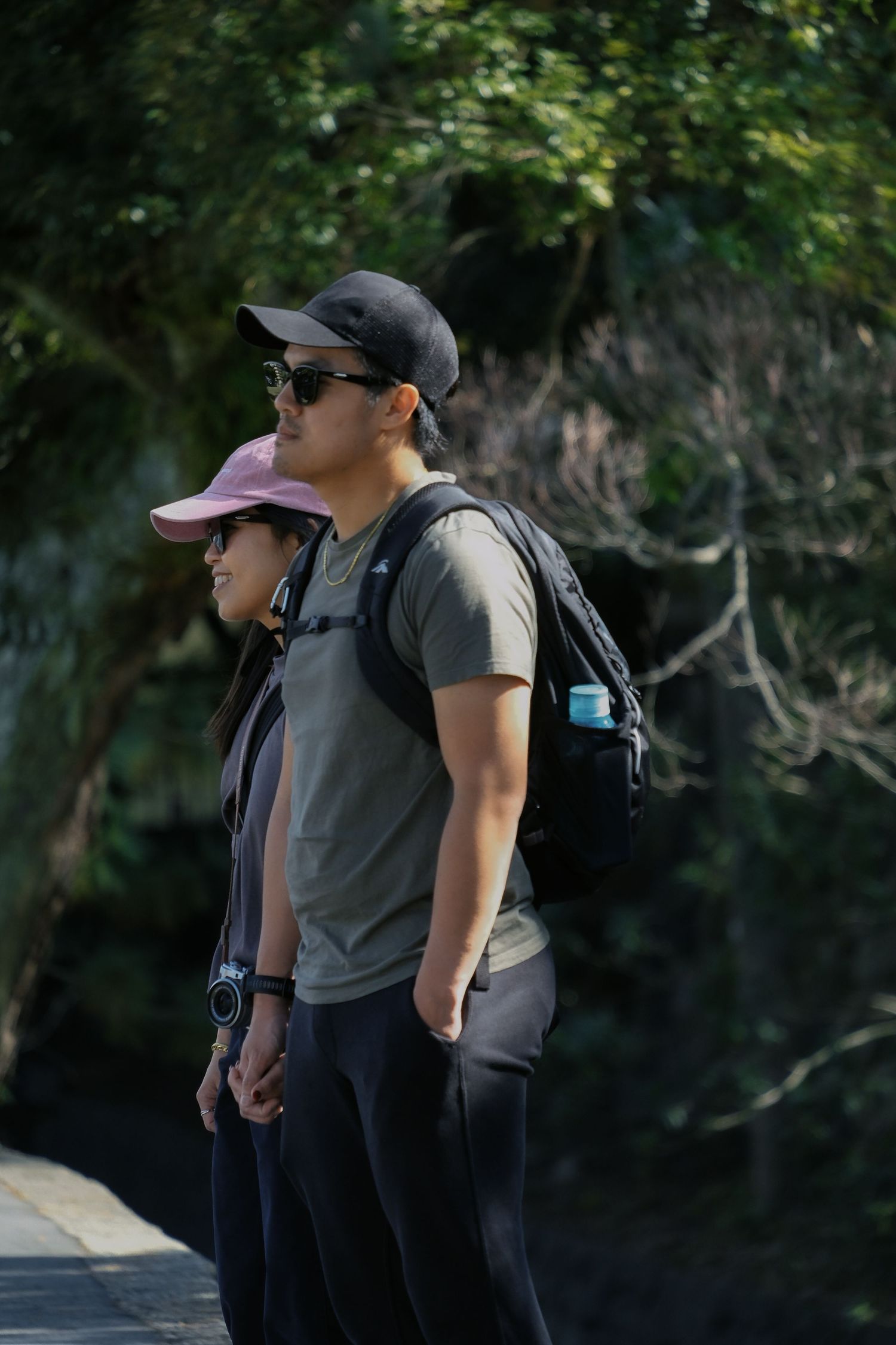 Couple wearing caps and sunglasses hiking together on a shaded forest trail, man carrying a backpack.