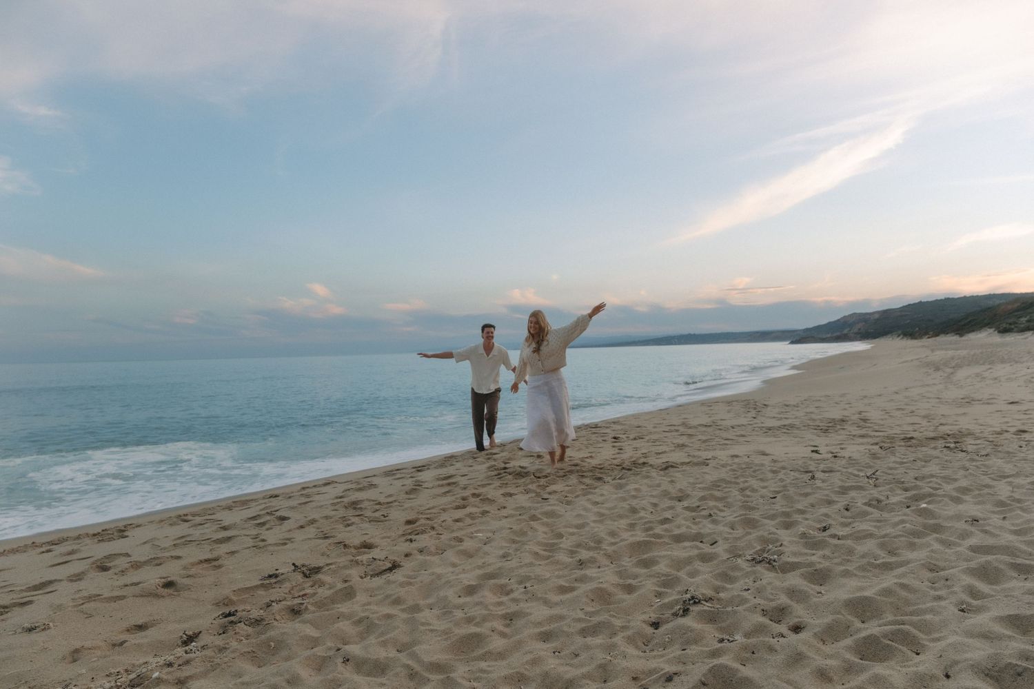 Couple walking hand-in-hand on a sandy beach at sunset, with ocean waves and a pastel sky in the background.