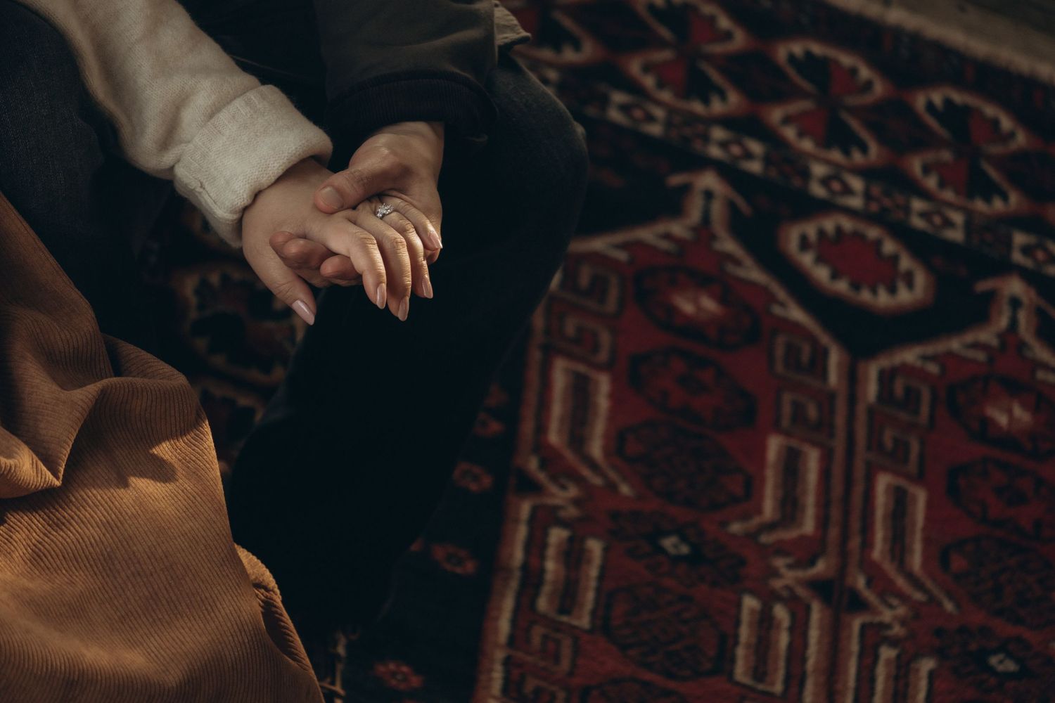 Close-up of hands with a ring resting on knees near a red patterned rug.