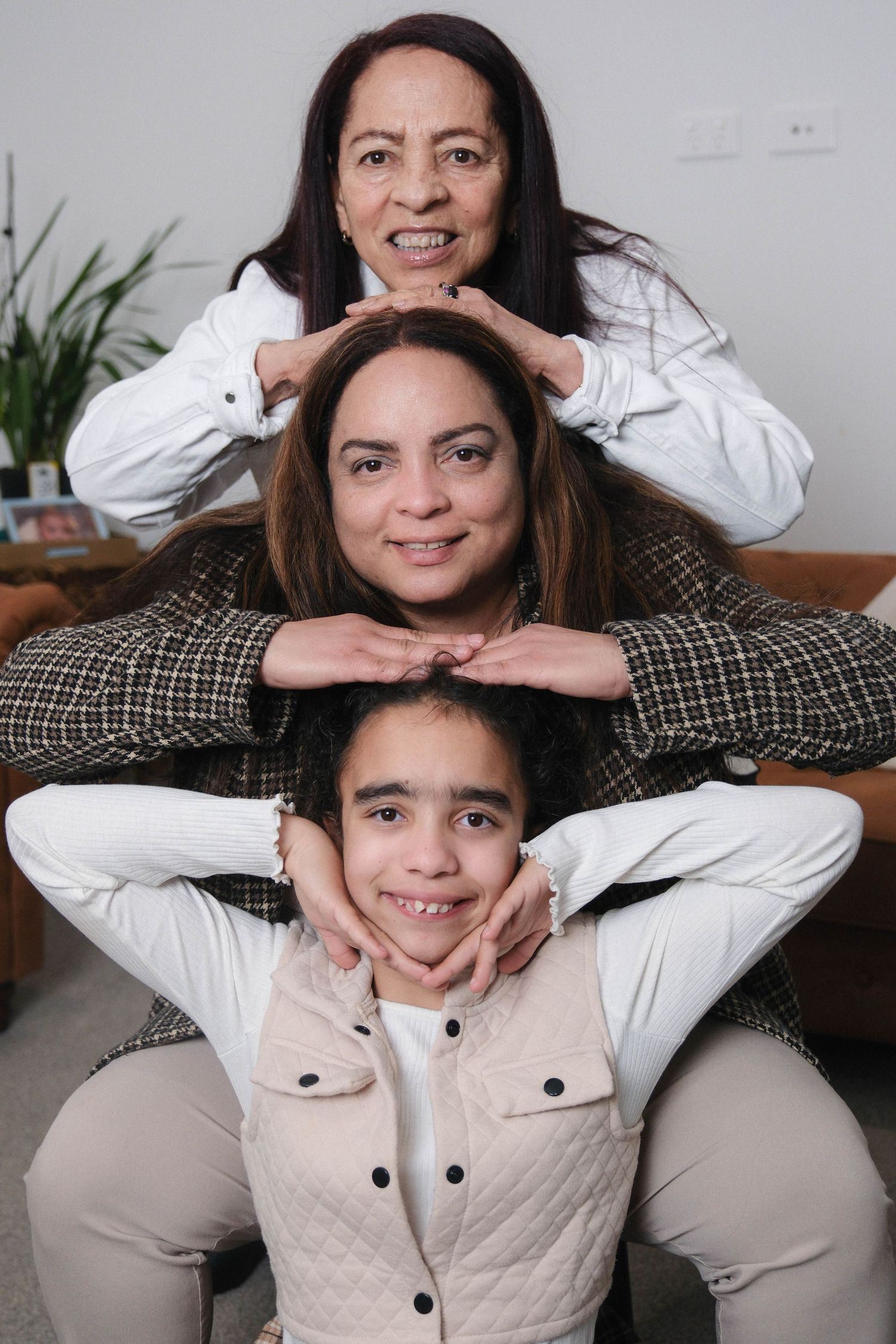 Three generations of women pose playfully stacked together, smiling warmly in a cozy living room setting.