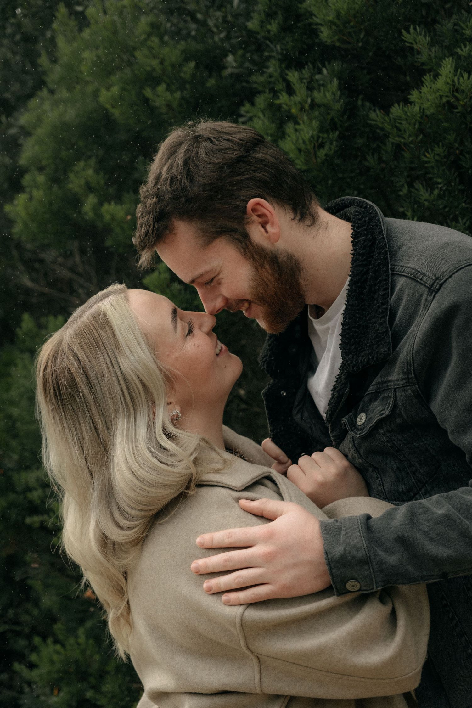 Happy couple nearly kissing outdoors, woman in beige coat and man in denim jacket among green trees.