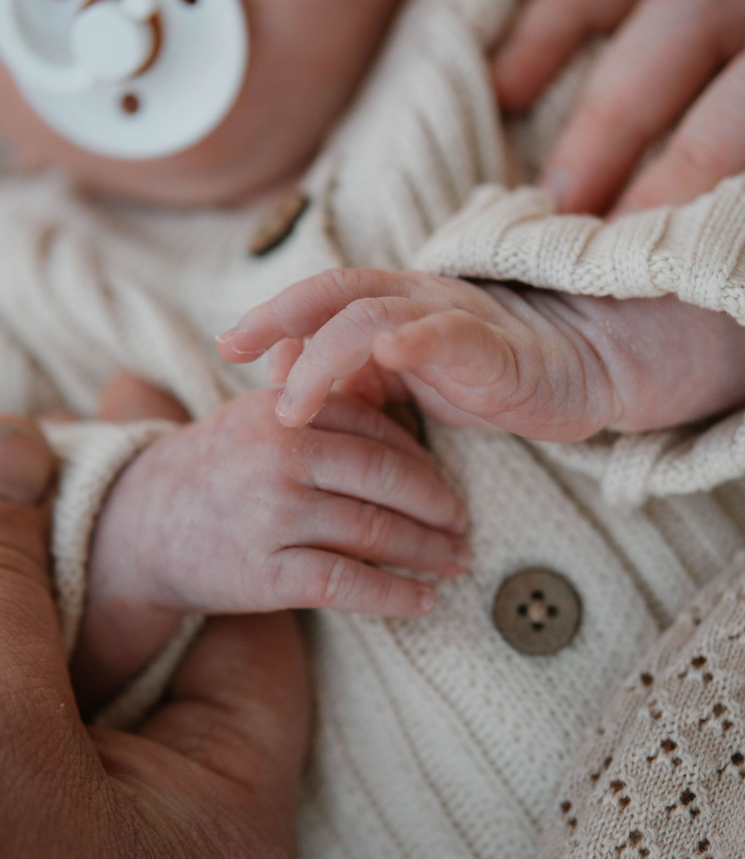Close-up of a newborn baby's tiny hand resting on a cream knit outfit with a pacifier visible.