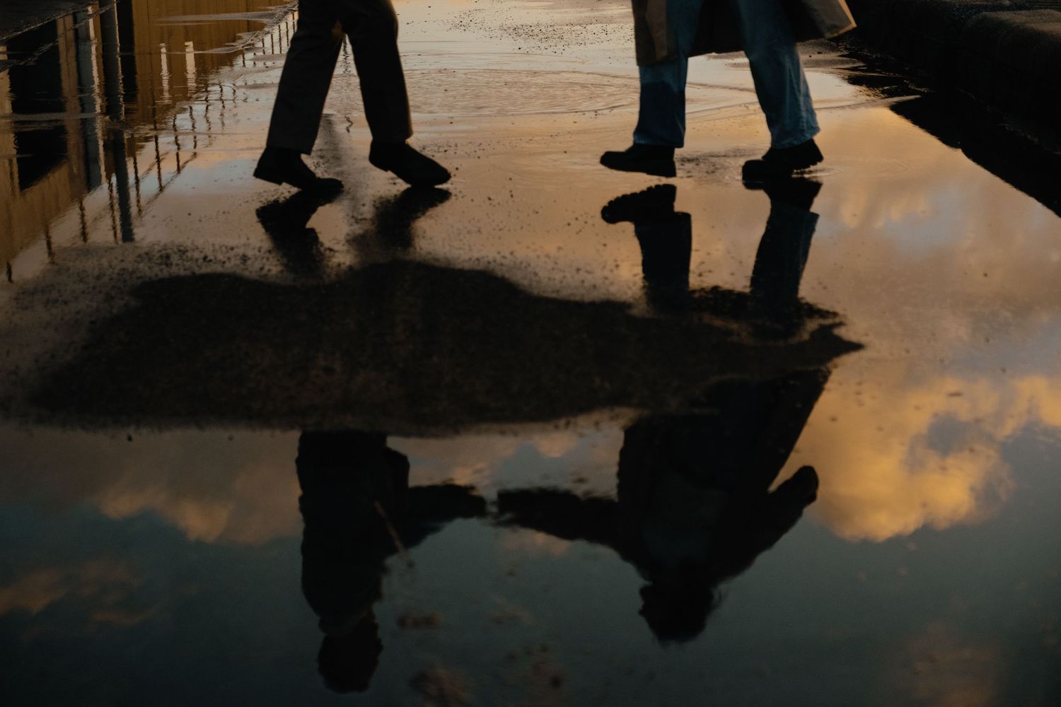 Two people walking on wet pavement with reflections of clouds and figures visible in a puddle at dusk.