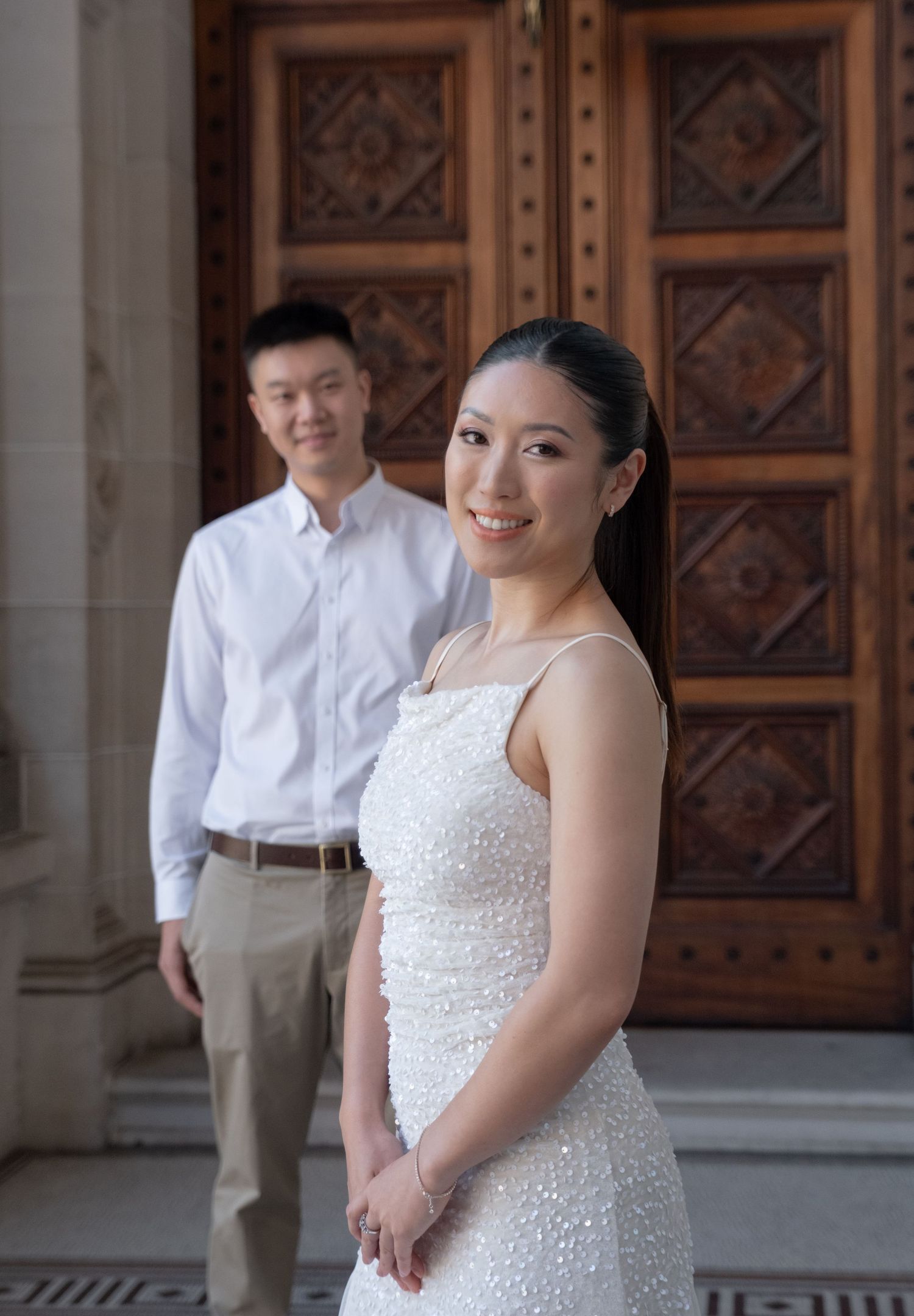 A bride in a white beaded gown smiles while her groom stands behind her before ornate wooden doors.