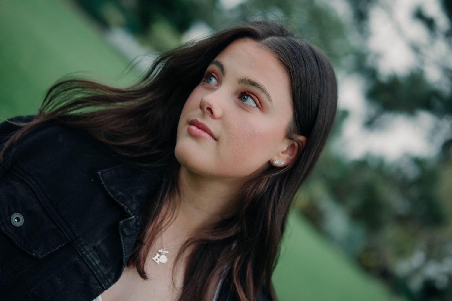 Young brunette woman in a black jacket gazing upward outdoors with soft green bokeh background.