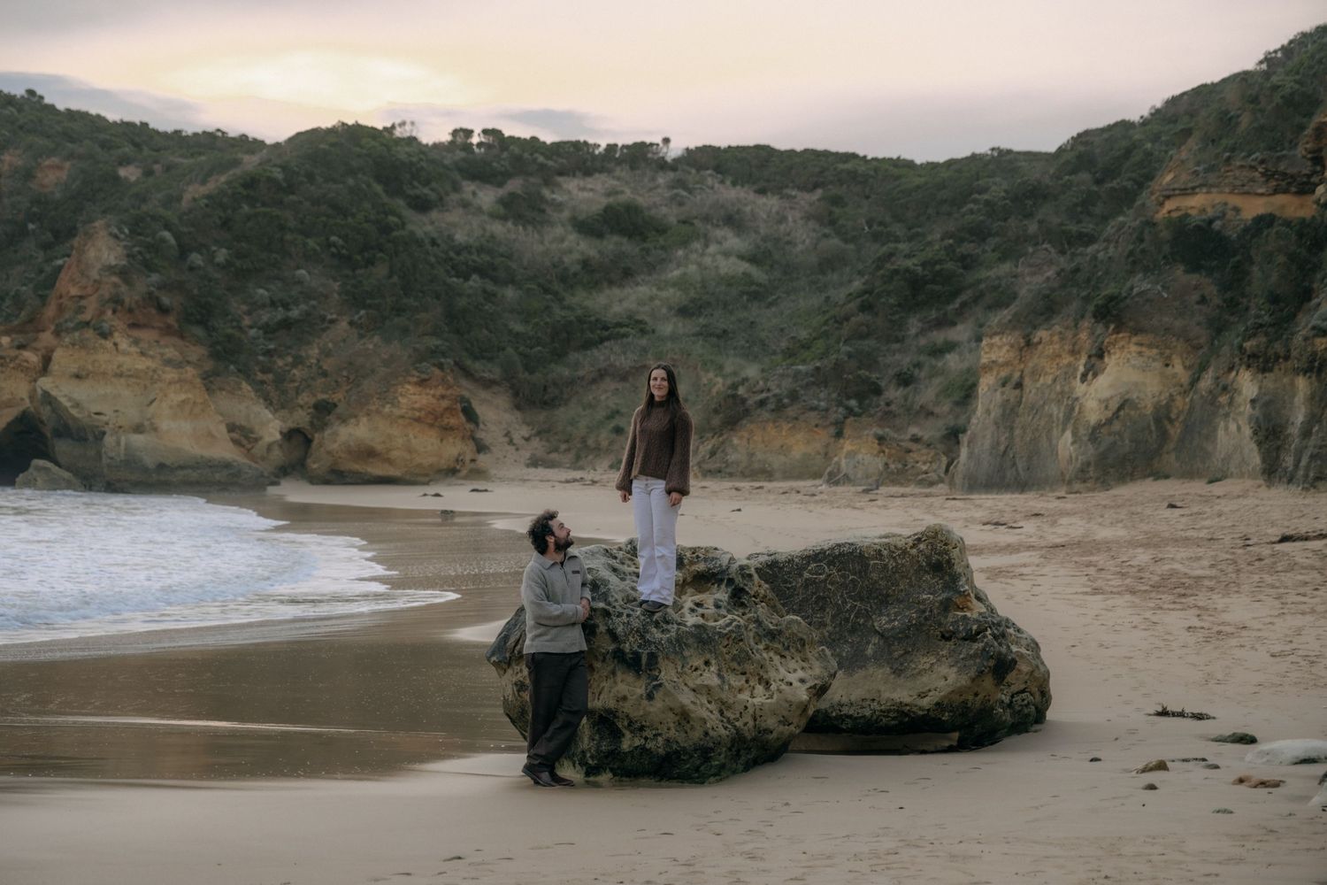 Couple on a scenic beach with rocky cliffs, one person standing on a boulder at sunset. Childers Cove near Warrnambool.