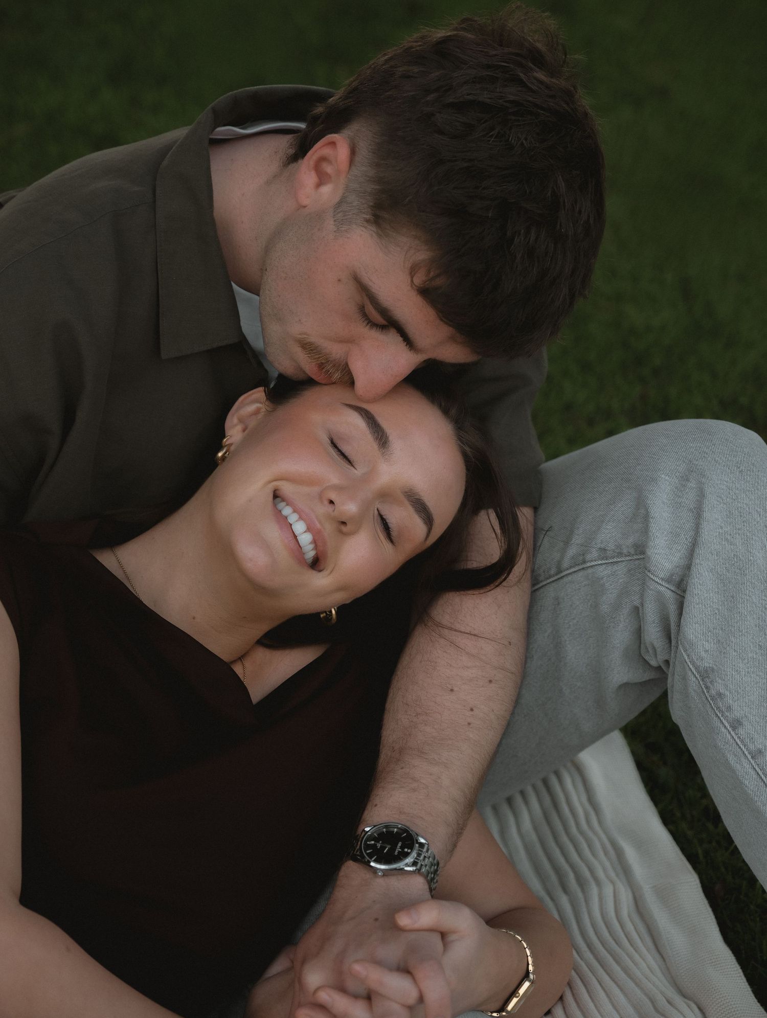 Young couple lying on grass, smiling and holding hands as man kisses woman on the forehead tenderly.