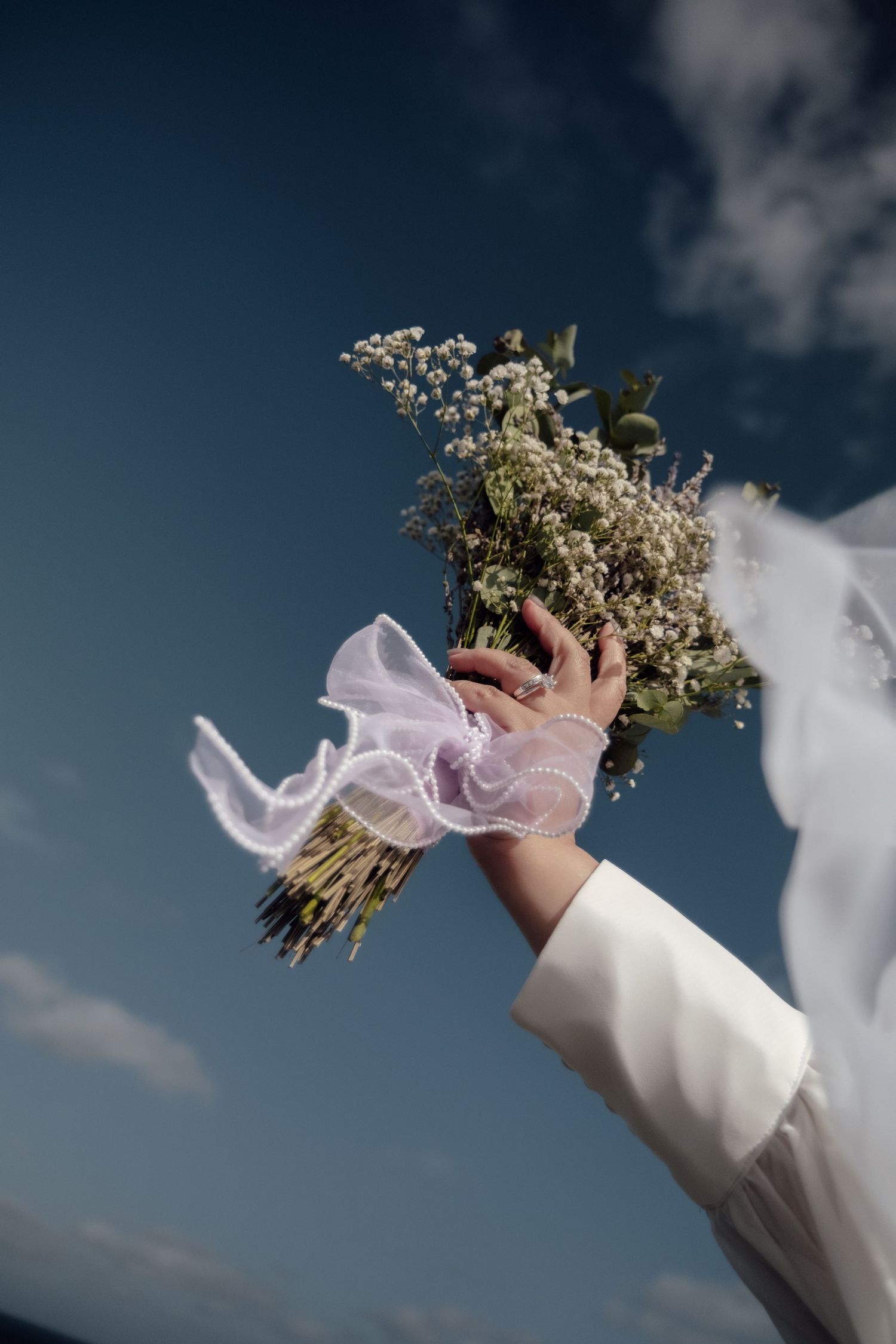 Bride holding a wildflower bouquet with pink ribbon raised against a blue sky, wearing a white dress with veil flowing.