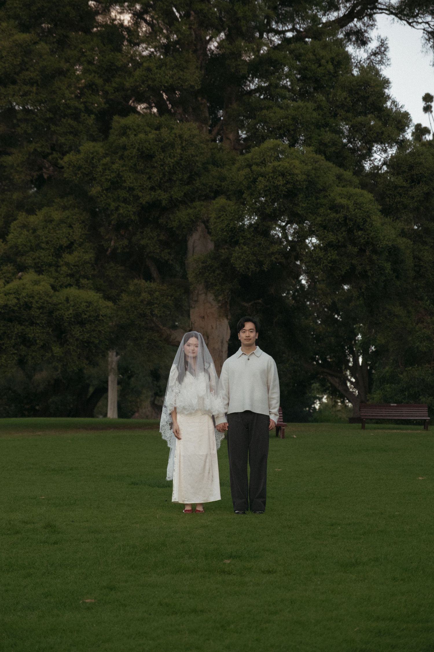 Couple in wedding attire holding hands on green lawn with large trees in background.