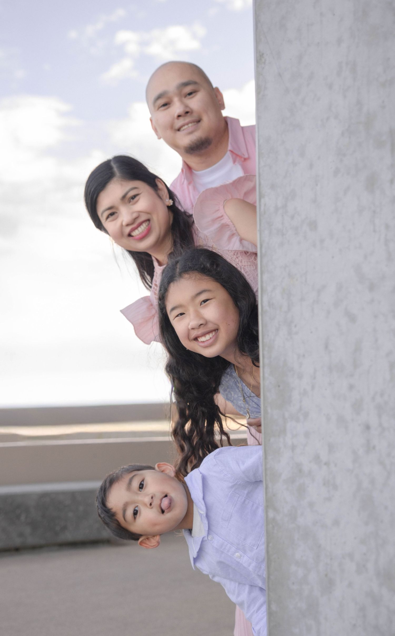 Smiling Asian family of four playfully peeking around a concrete column with ocean backdrop.