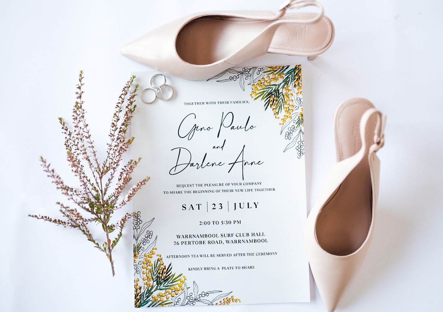 Wedding invitation with floral design beside blush heels and rings on white background.