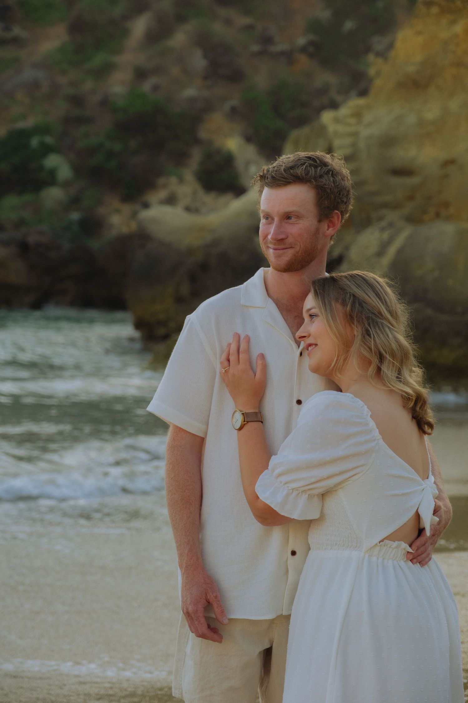 Romantic couple in white outfits embracing on a beach with dramatic rocky cliffs in the background.