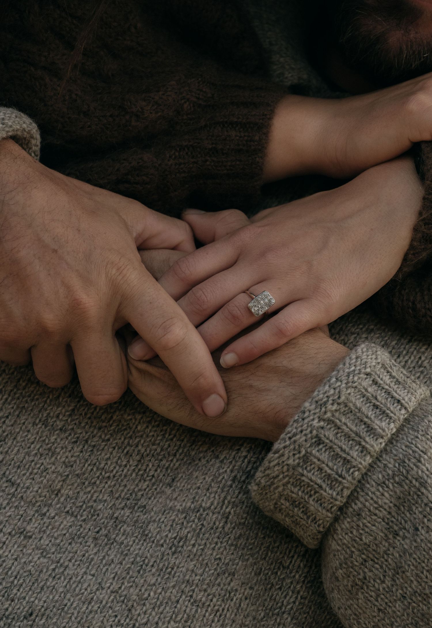 Close-up of couple holding hands showing diamond engagement ring, wearing cozy knit sweaters.