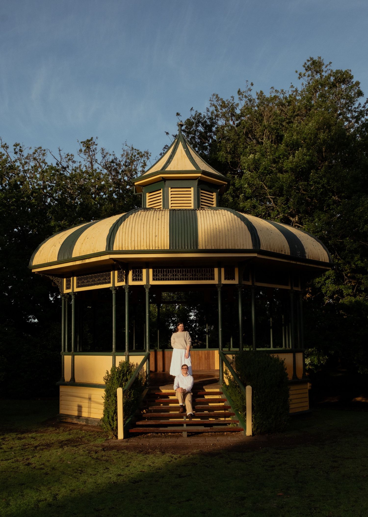 Couple in white outfits descending steps of a charming vintage gazebo at golden hour. Warrnambool Botanic Gardens.