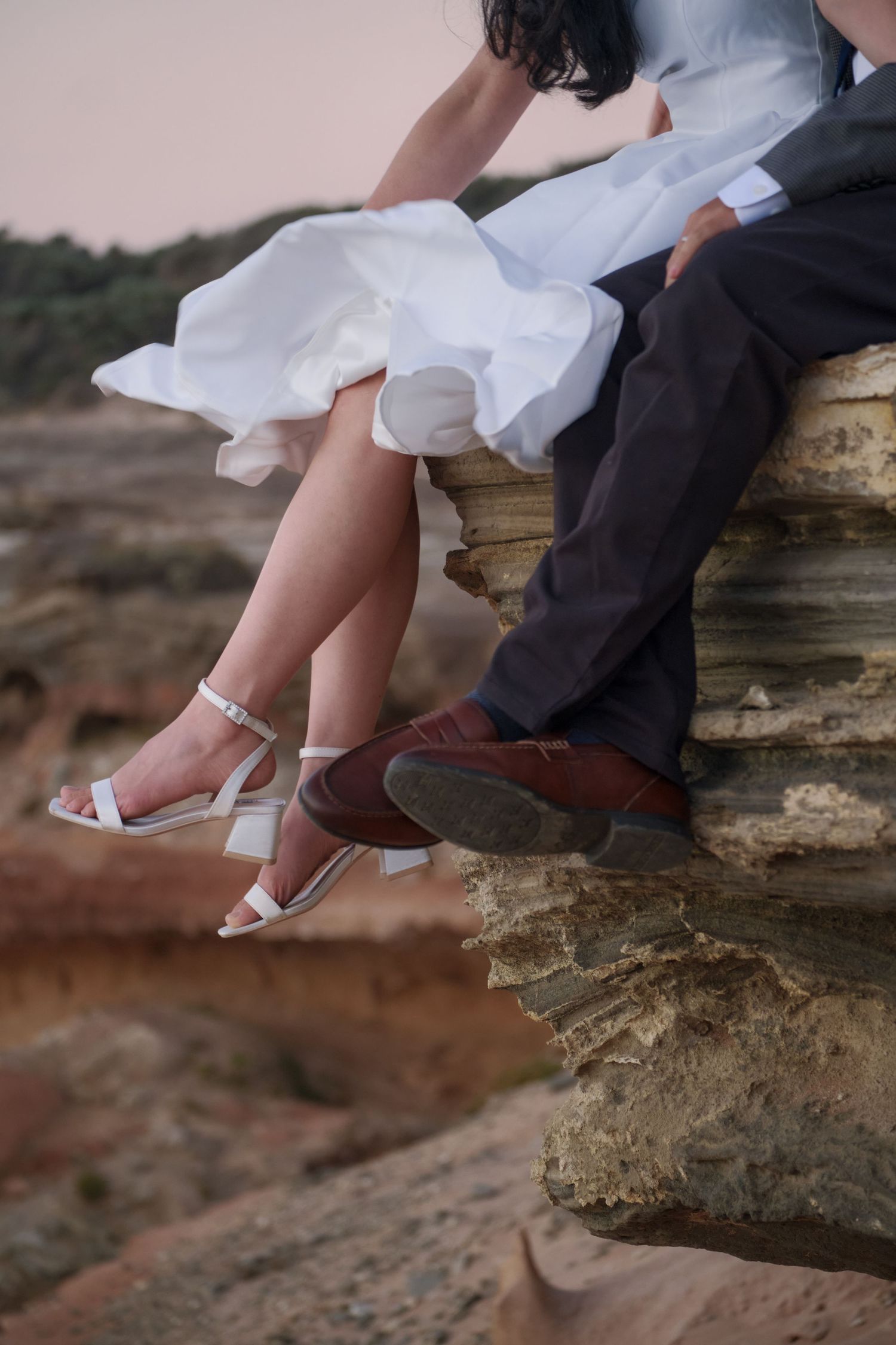 Close-up of bride in white dress and heels sitting beside groom in dark pants and brown shoes on rocky ledge.