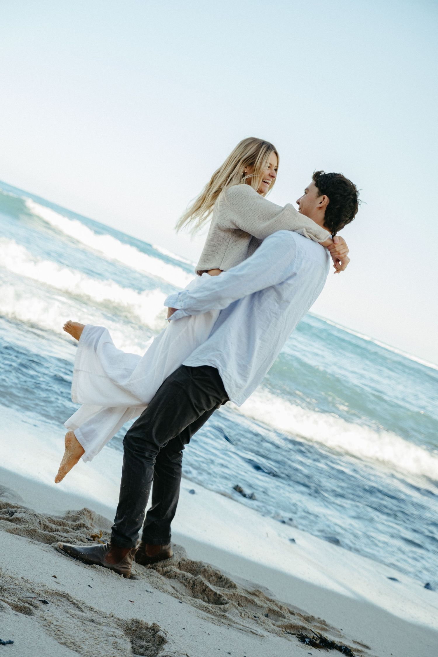 A couple shares a romantic moment on the beach, with the man lifting the woman in his arms near ocean waves.